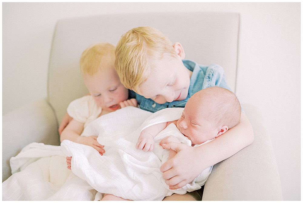 Red-Haired Toddler Boy And Girl Hold And Smile At Their Newborn Baby Brother.