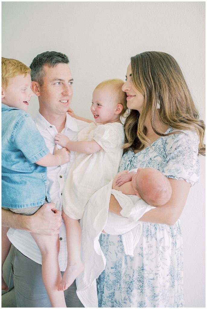 Mother And Father Hold Their Two Toddler And Newborn Baby And Smile At One Another During Their Joint Base Andrews Newborn Session.