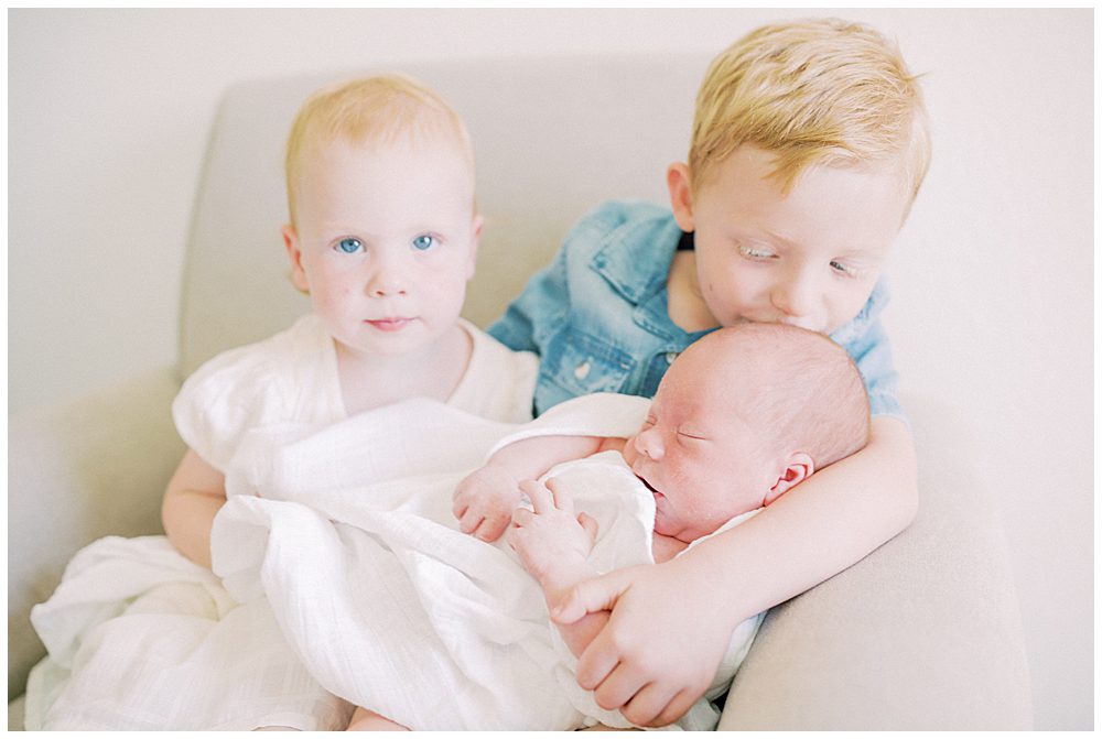 Toddler Boy Sits With His Toddler Sister And Kisses Their Baby Brother Brother On The Head.