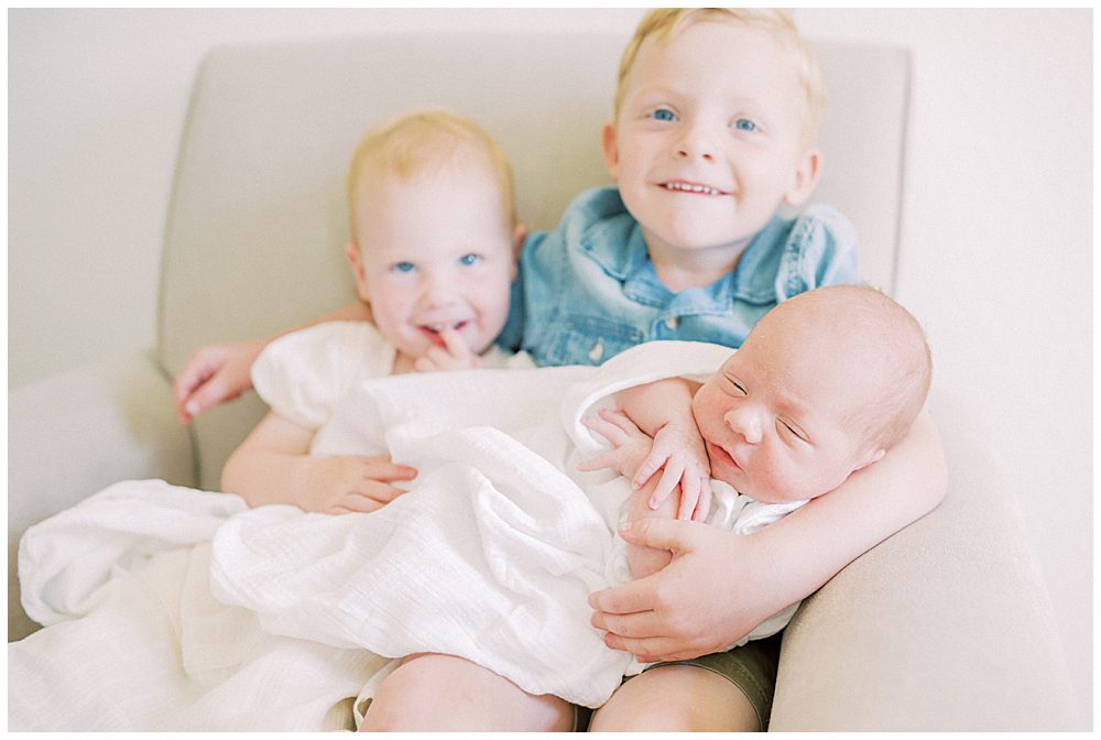 Toddler Boy And Girl Smile As They Hold Their Newborn Baby Brother During Their Joint Base Andrews Newborn Session.