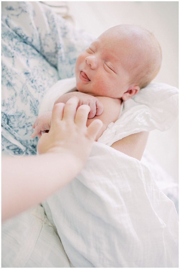 Newborn Baby Smiles While Held By His Mother And Older Sibling Touches His Hand.