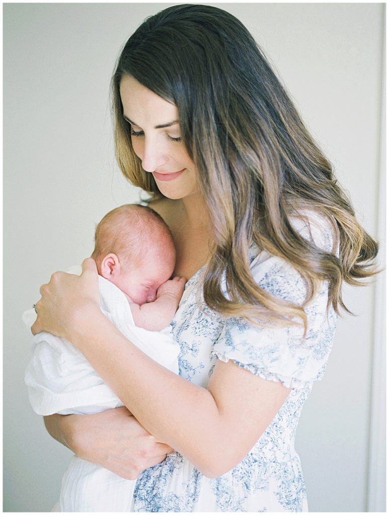 Mother With Brown Hair Smiles As She Holds Her Newborn Baby Up To Her Chest During Her Joint Base Andrews Newborn Session.