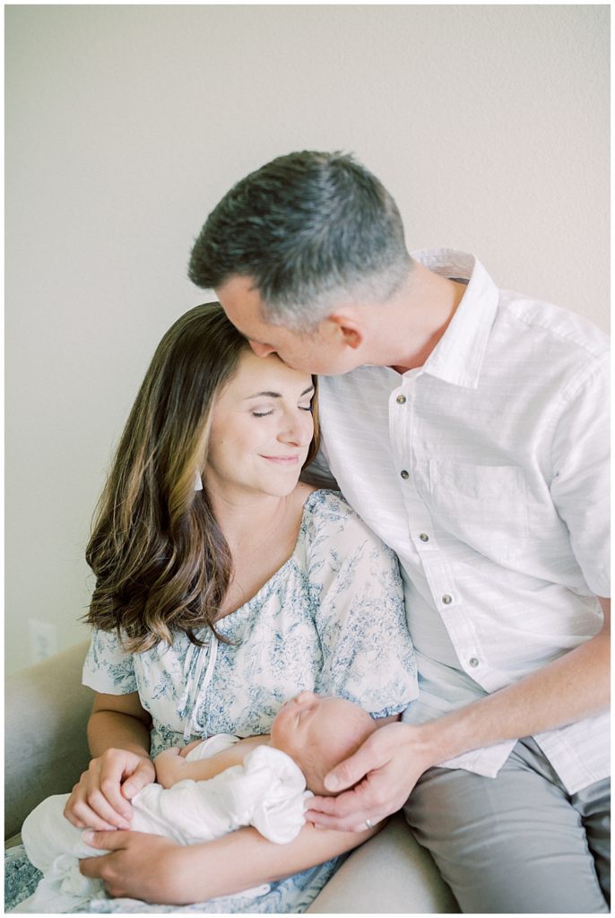 Father Kisses His Wife's Forehead As She Smiles, Holding Their Newborn Baby During Their Joint Base Andrews Newborn Session.