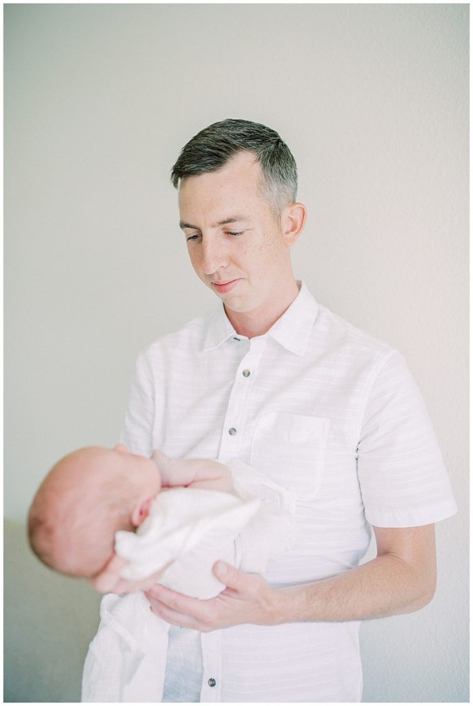 Father Holds His Newborn Baby In Front Of Him, Looking Down At Him.