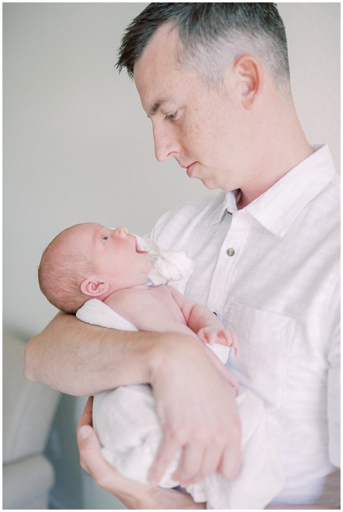 Father Holds His Newborn, Looking Down At Him While The Baby Looks Back At Him With An Open Mouth.