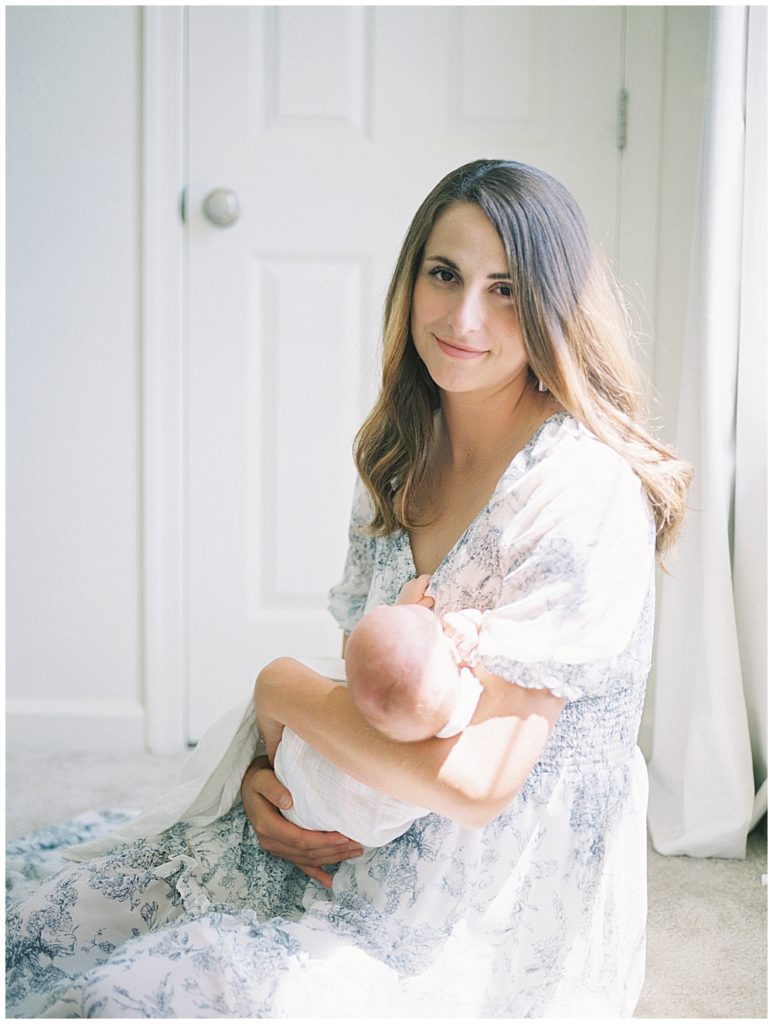 Mother Sits In Front Of A Sun-Soaked Window, Holding Her Newborn Baby And Looking Directly At The Camera.