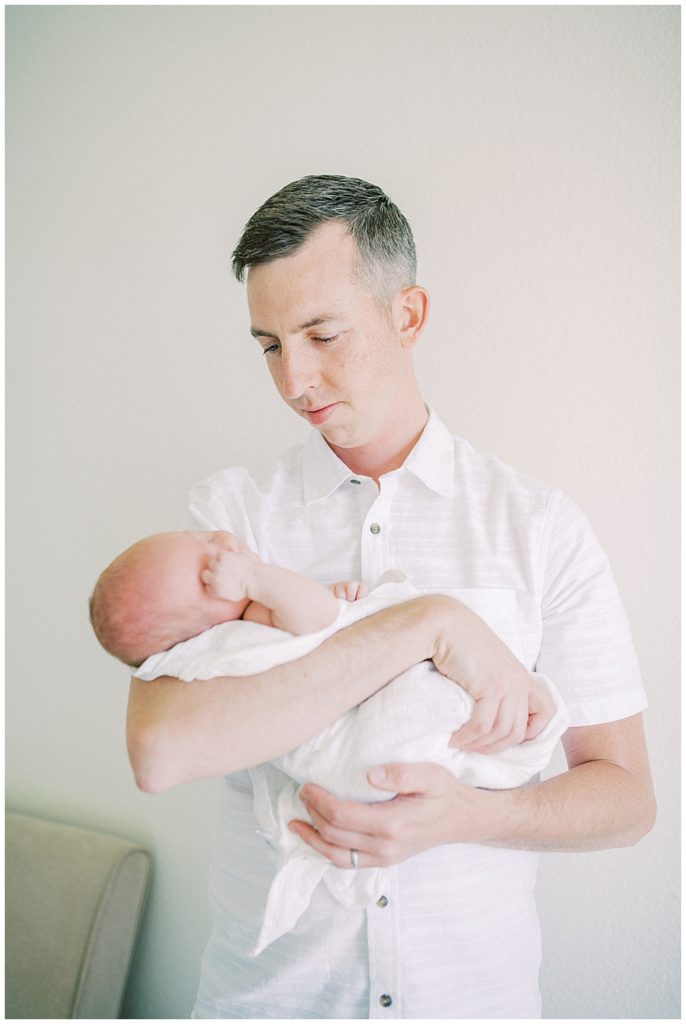 A Father Looks Down At His Newborn Baby While Holding Him. 