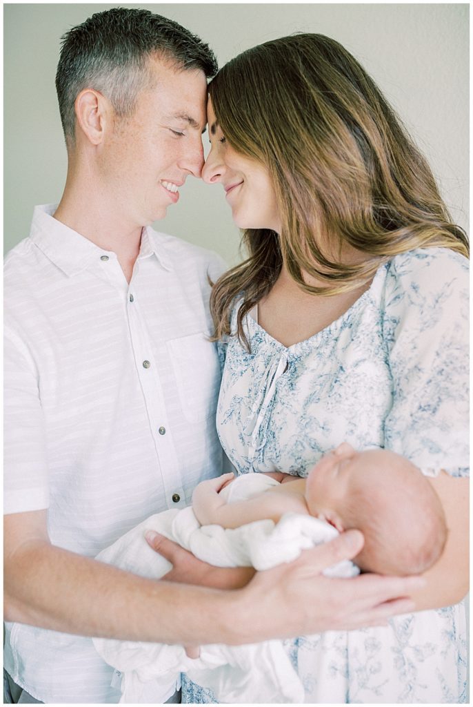 Father And Mother Smile And Lean In Together While Holding Their Baby During Their Joint Base Andrews Newborn Session.