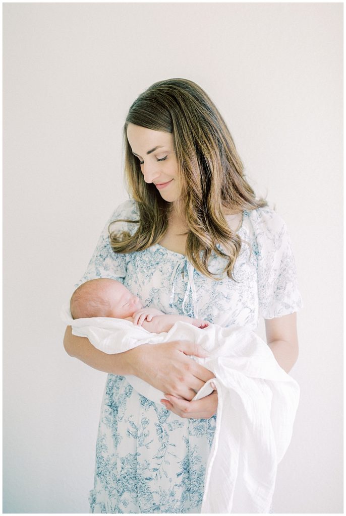 New Mother Smiles Down At Her Newborn Baby Wrapped In A White Swaddle.