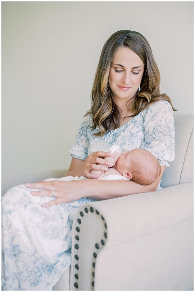 New Mother In Blue And White Dress Sits In A Chair And Gives Her Newborn Baby A Bottle During Her Joint Base Andrews Newborn Session.