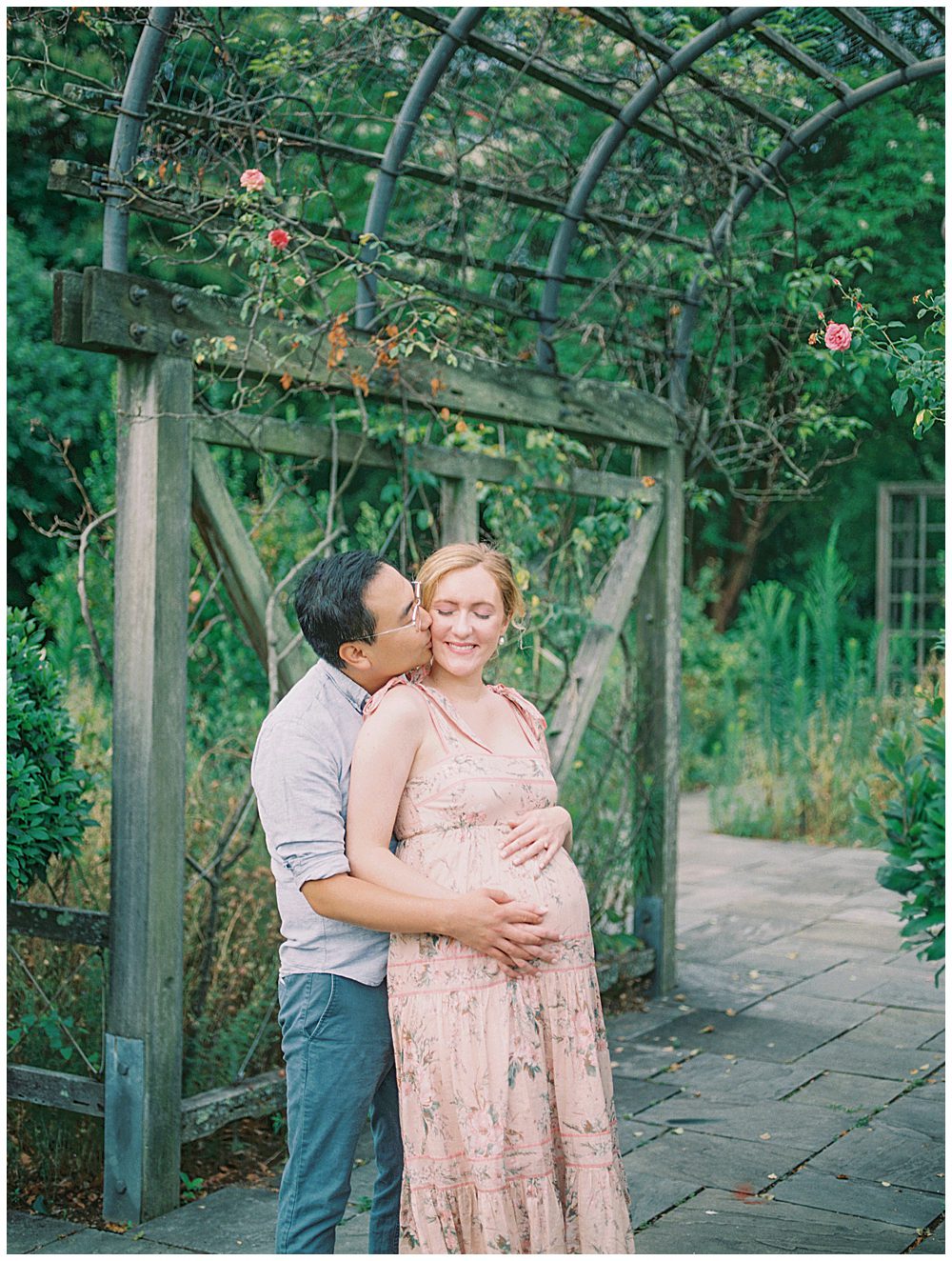 Husband Leans Over To Kiss His Wife's Cheek While They Stand Under A Floral Arch During Their Maternity Session At The National Arboretum.