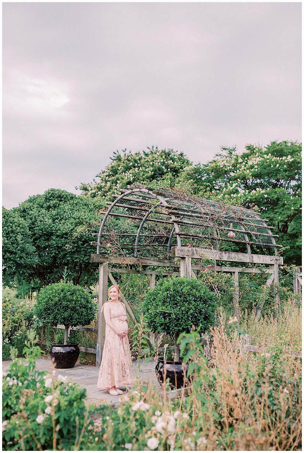 Blonde Pregnant Mother Stands Under Floral Arch During Maternity Session In Summer.