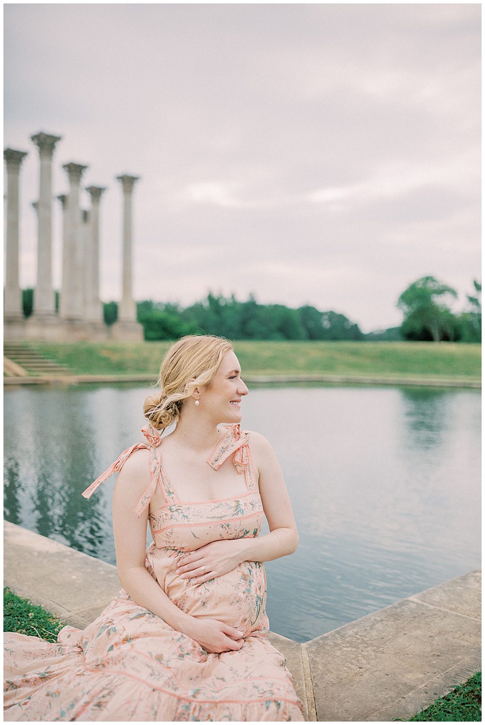 Blonde Mother In Zimmerman Pink Dress Sits In Front Of Pond With Hands On Her Belly During Her Maternity Session.