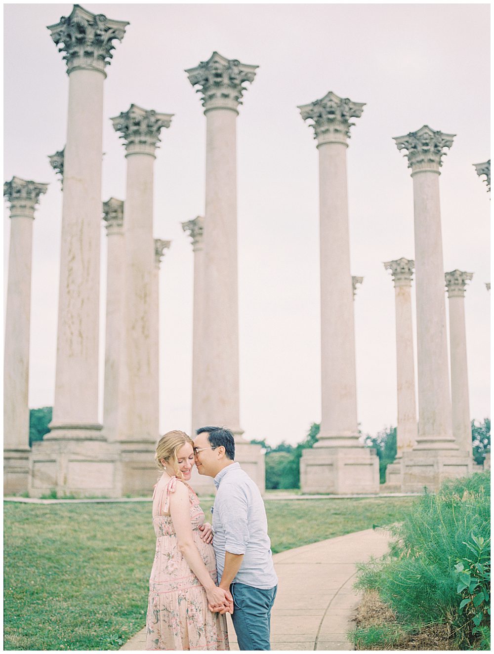 Husband Leans Over To Nuzzle His Wife's Cheek While They Stand On The Sidewalk In Front Of The National Arboretum Columns.