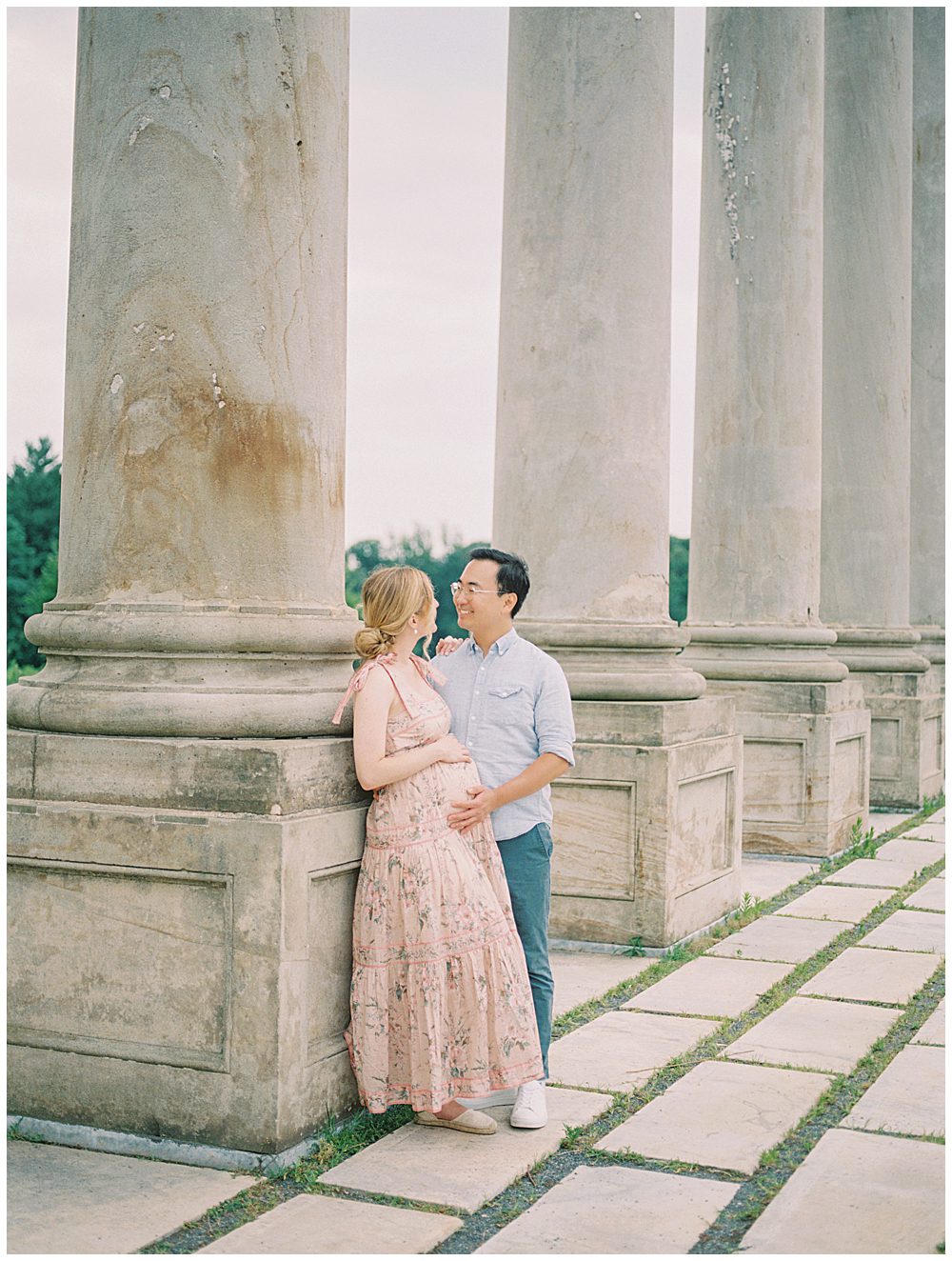 Mother In Pink Zimmerman Dress Leans Against Pillar At The National Arboretum As Her Husband Places A Hand On Her Pregnant Belly.