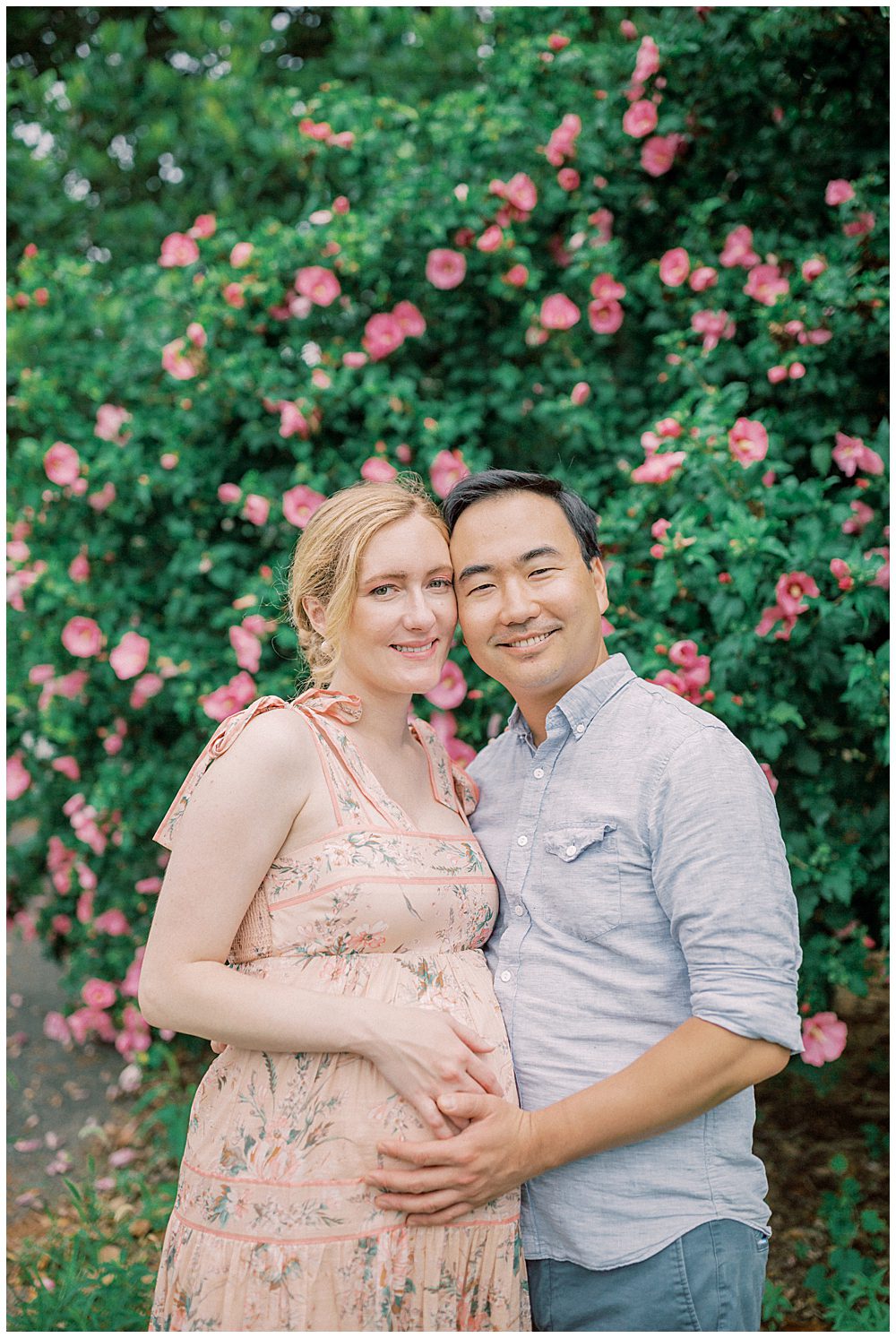 Expecting Parents Lean Into One Another And Smile While Standing In Front Of A Rose Bush.