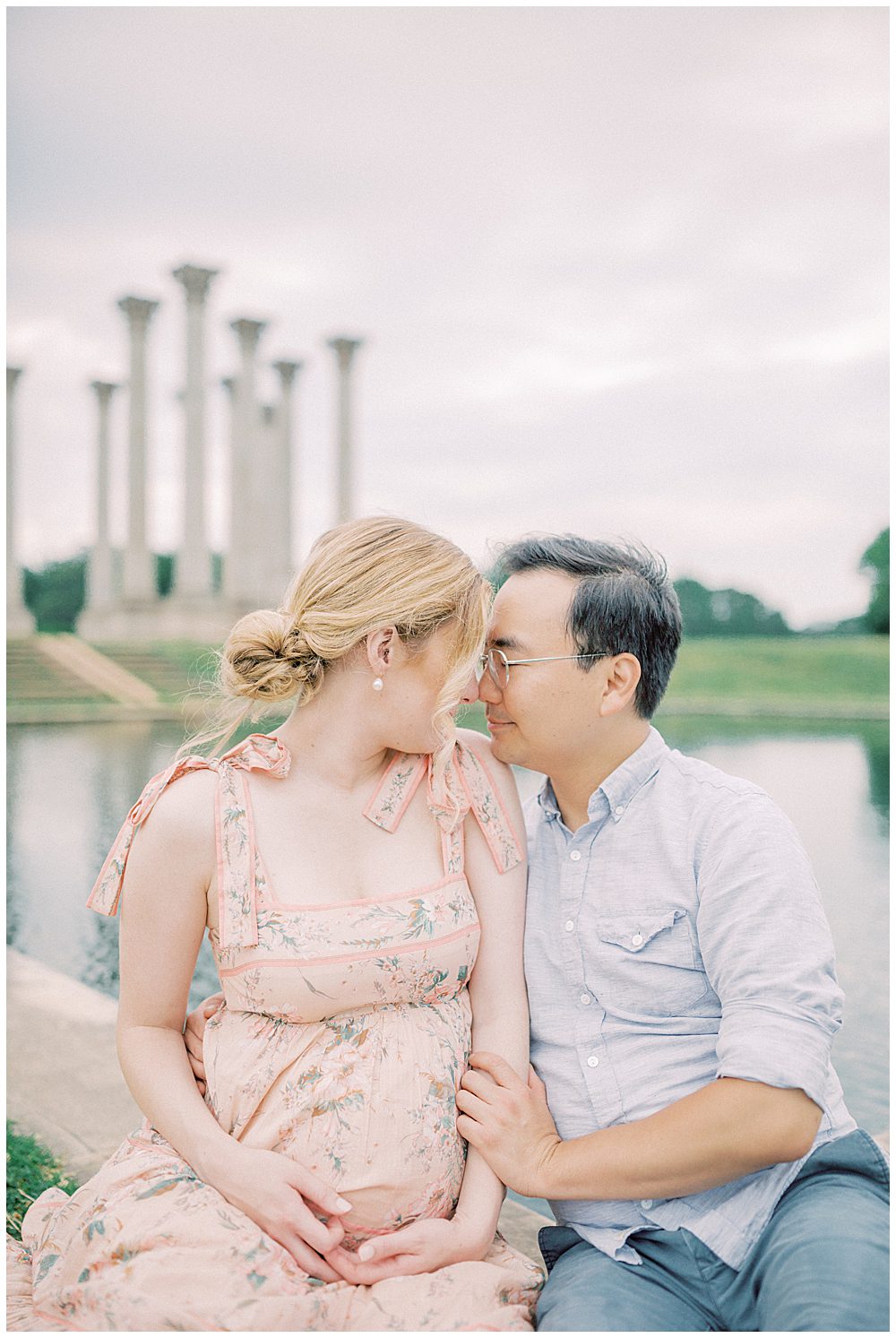 Husband And Wife Sit Together And Lean Into One Another In Front Of The Pond At The National Arboretum During Their Maternity Session.