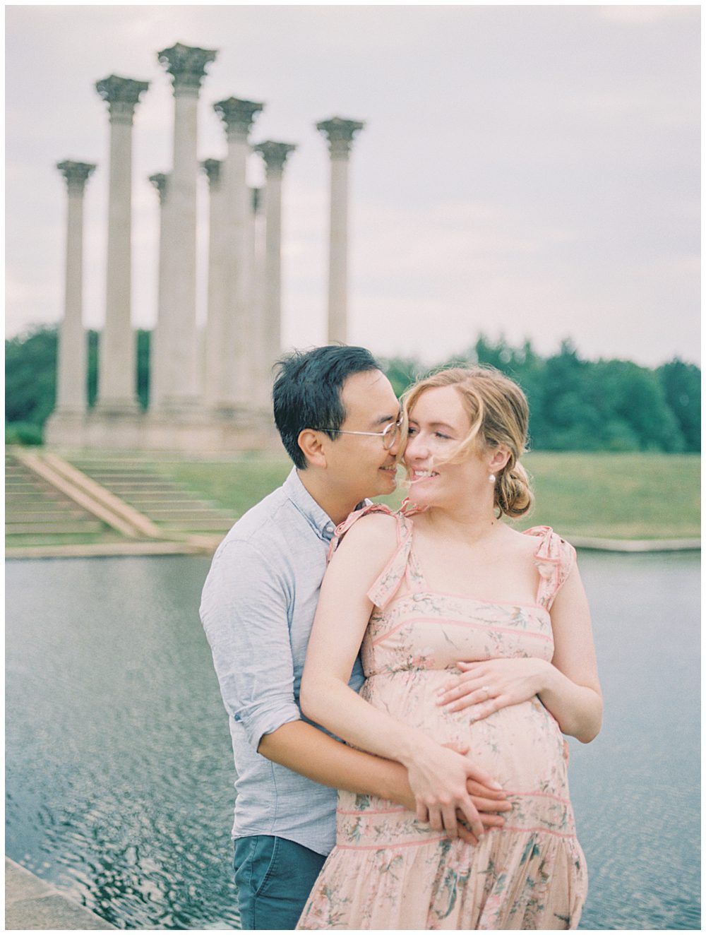 Husband Leans Into Wife As They Stand In Front Of Pond At The National Arboretum During Their Maternity Session.