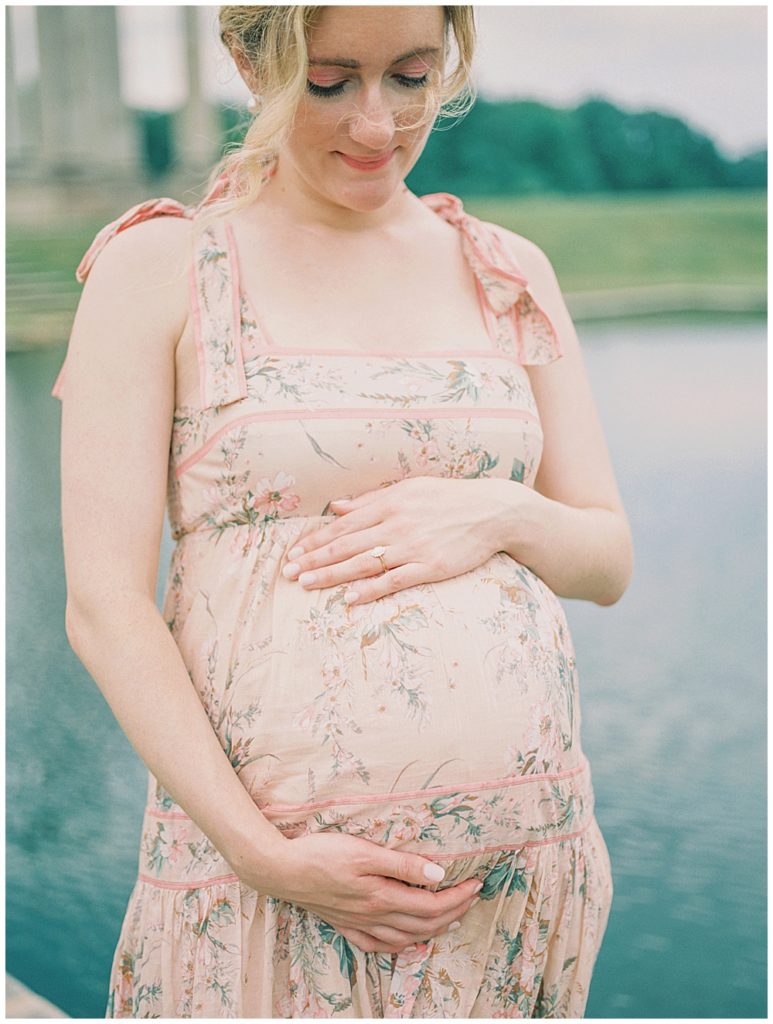 Mother Stands In Front Of A Blue Pond, With One Hand On Top And One Hand Below Her Pregnant Belly.