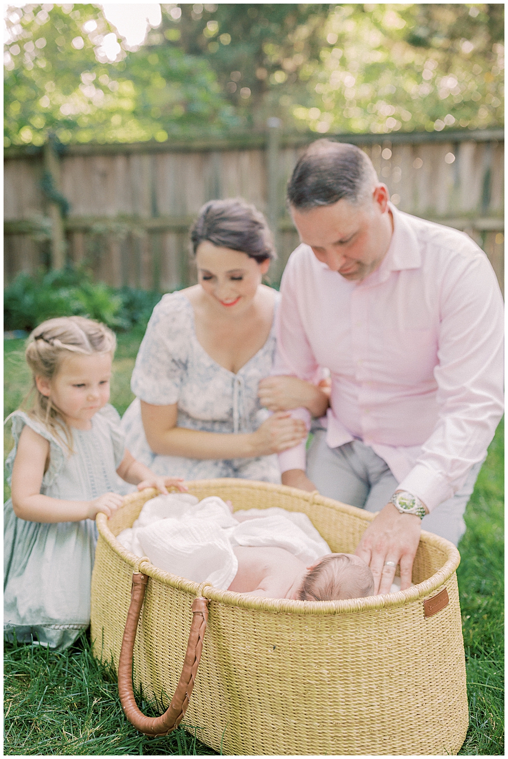 Mother, Father, And Big Sister Lean Over A Moses Basket Admiring Their New Baby During Their Outdoor Newborn Session