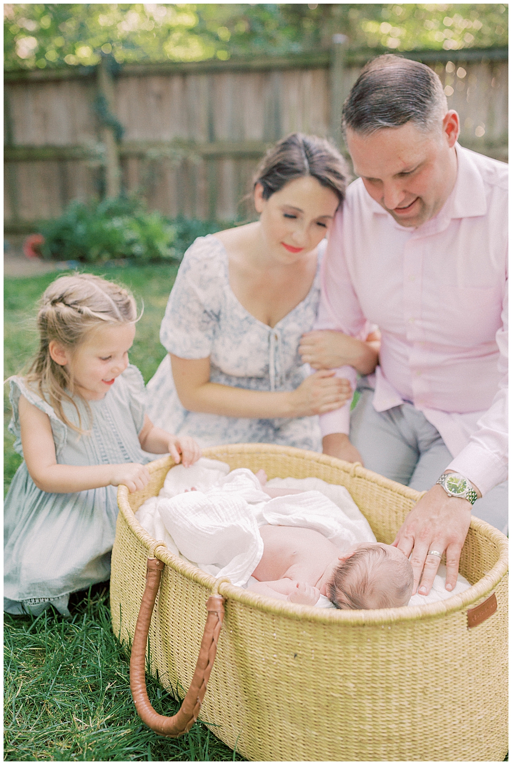 Mother, Father, And Toddler Sister Sit Outside And Look Over Moses Basket With Newborn Baby