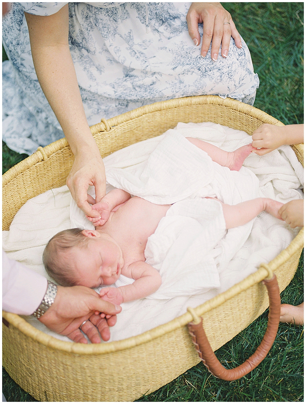 Newborn Baby Lays In Moses Basket While Family Hold Her Hands And Feet