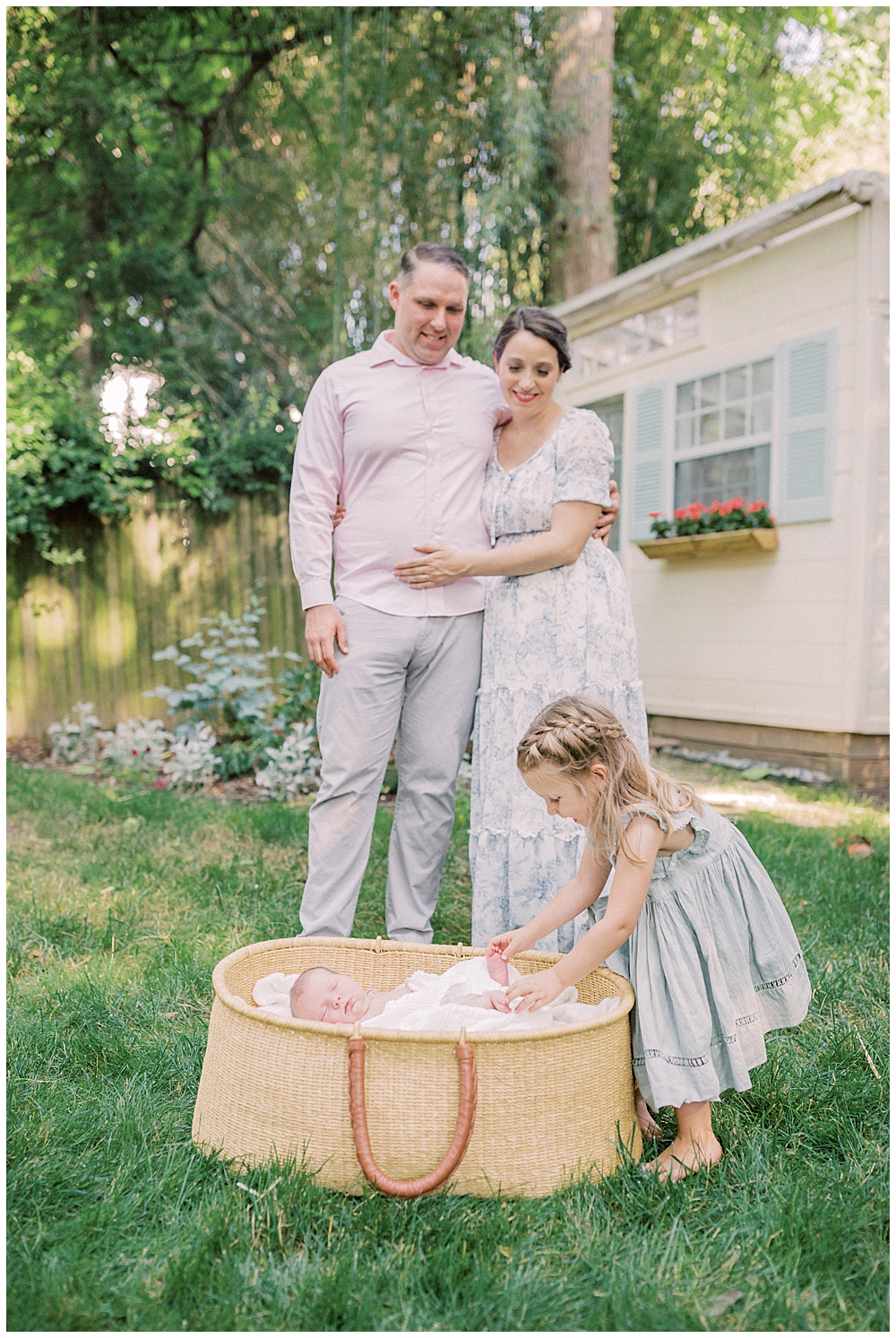 Parents Look Down As Their Toddler Girl Plays With Their Newborn Baby In A Moses Basket During Their Outdoor Studio Newborn Session