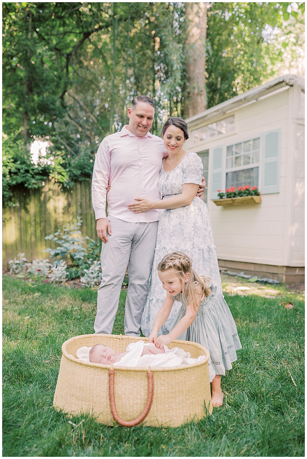 Mother And Father Smile Down At Their Toddler Daughter As She Leans Over A Moses Basket With Their Newborn Baby