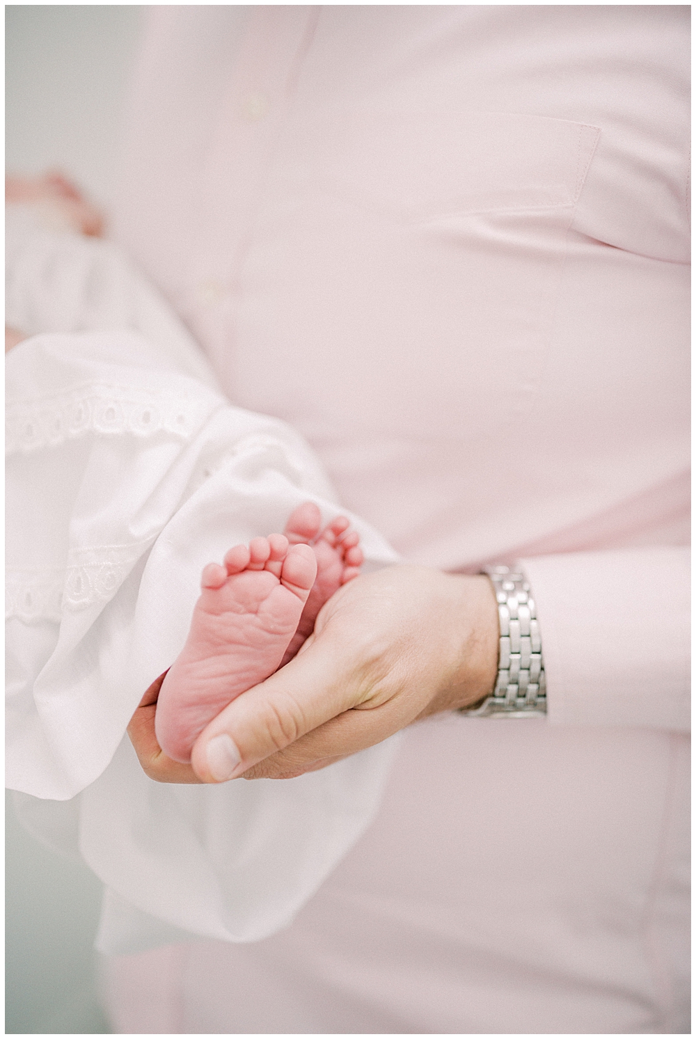 A Dad In A Pink Shirt Holds His Newborn's Feet In His Hands