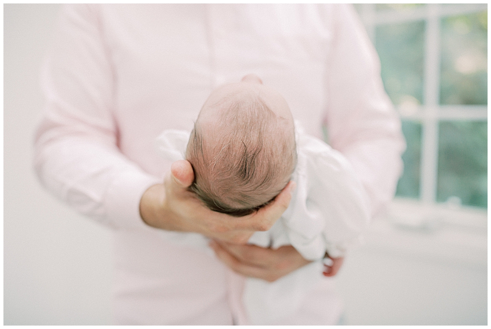 Close-Up Of Newborn Head While Newborn Is Held By Her Father In A Pink Shirt