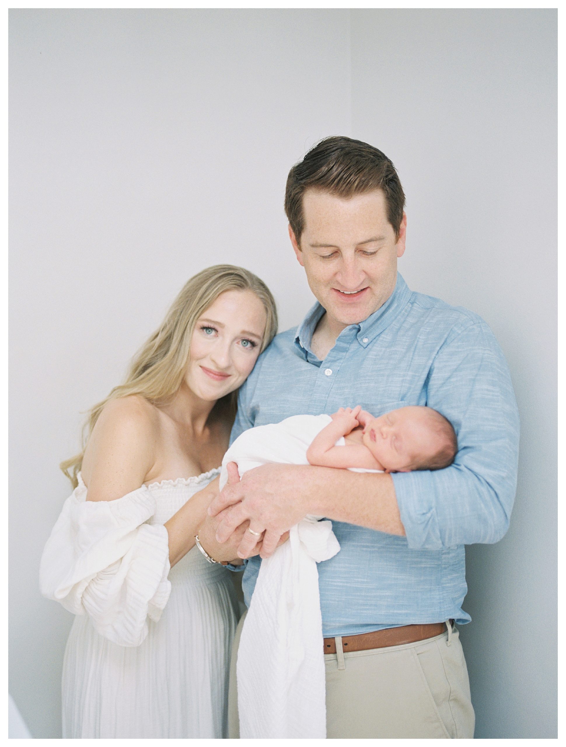 Blonde Mother Leans On Husband Who Holds And Looks Down At Newborn Son.