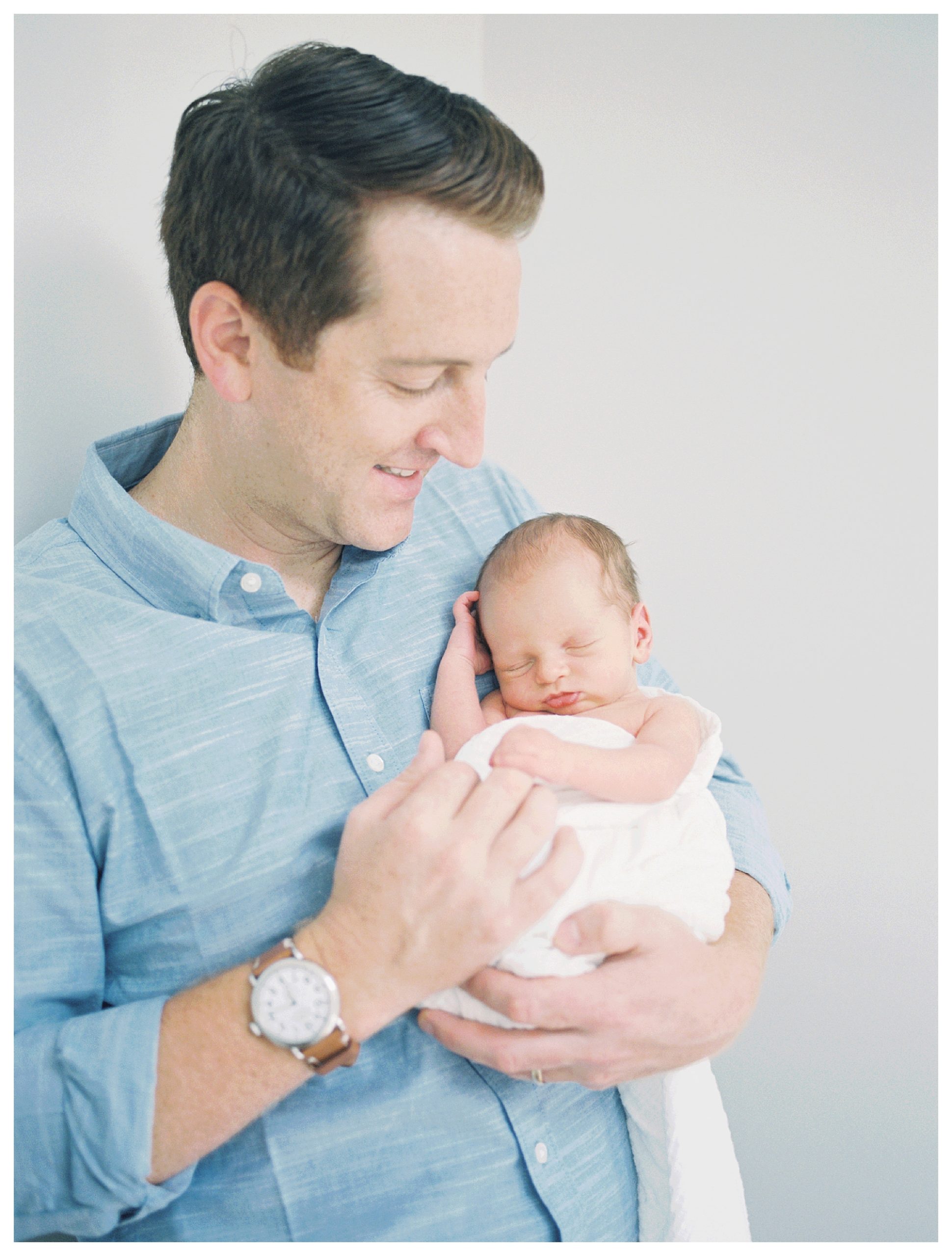 Father Holds Up Newborn Son To His Chest And Smiles Down At Him.