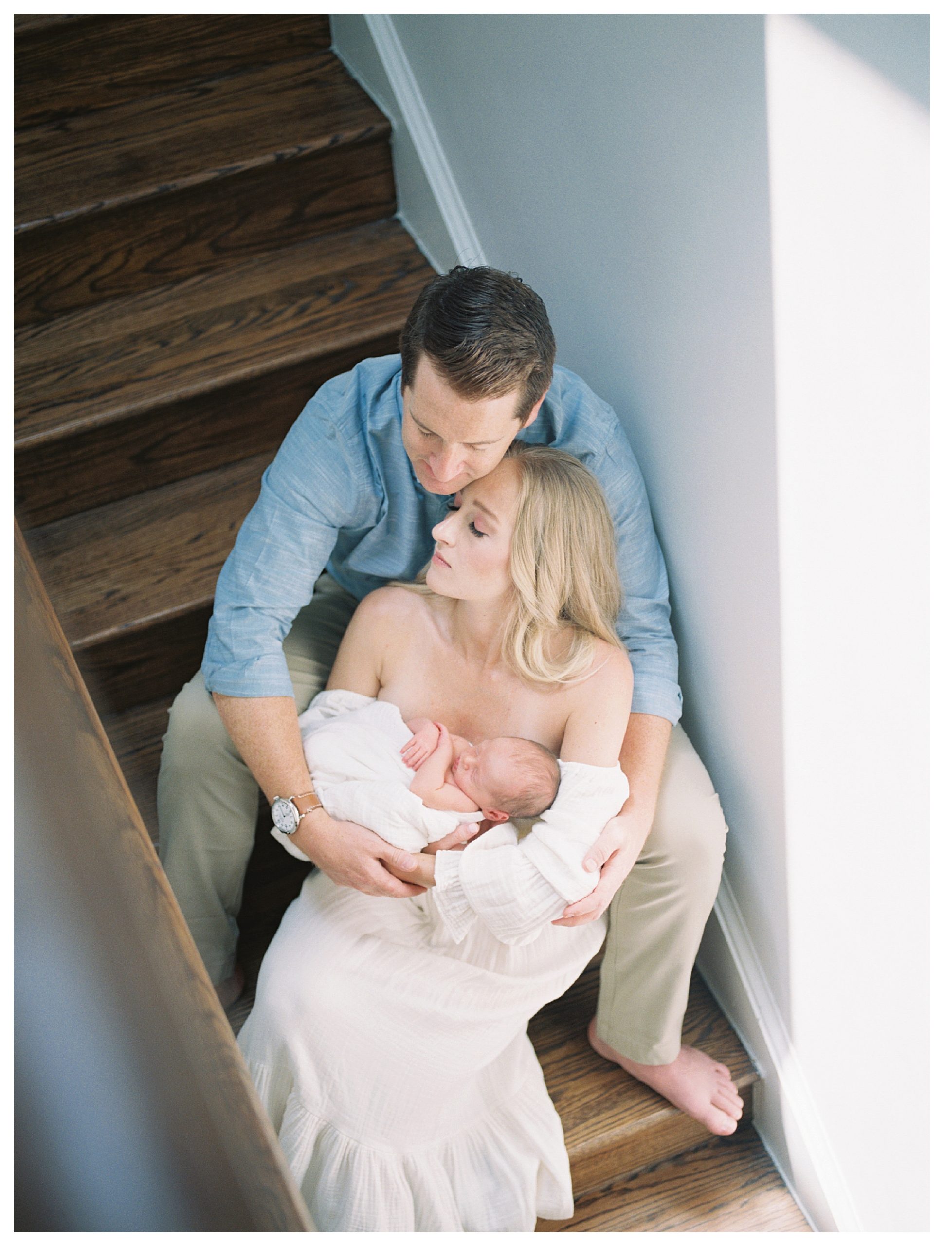 Father And Mother Sit On Steps, Leaning Into One Another As They Hold Their Newborn Baby.