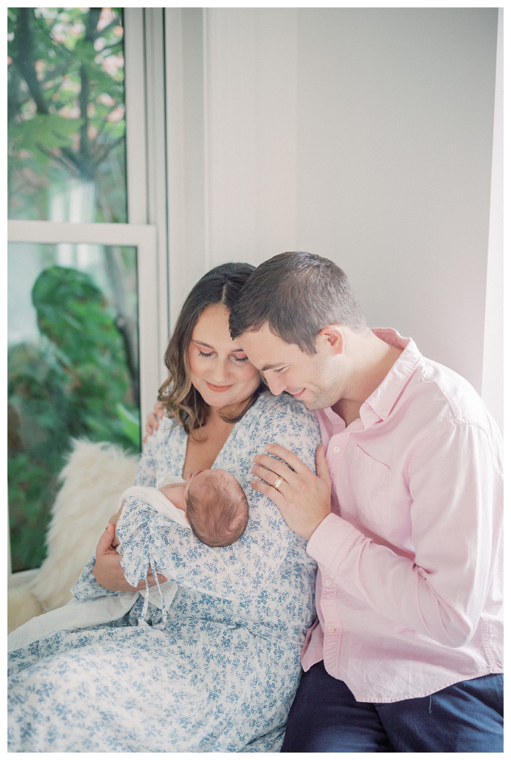 Father Leans Over His Wife's Shoulder And Smiles At Their Newborn Baby During In-Home Newborn Session.