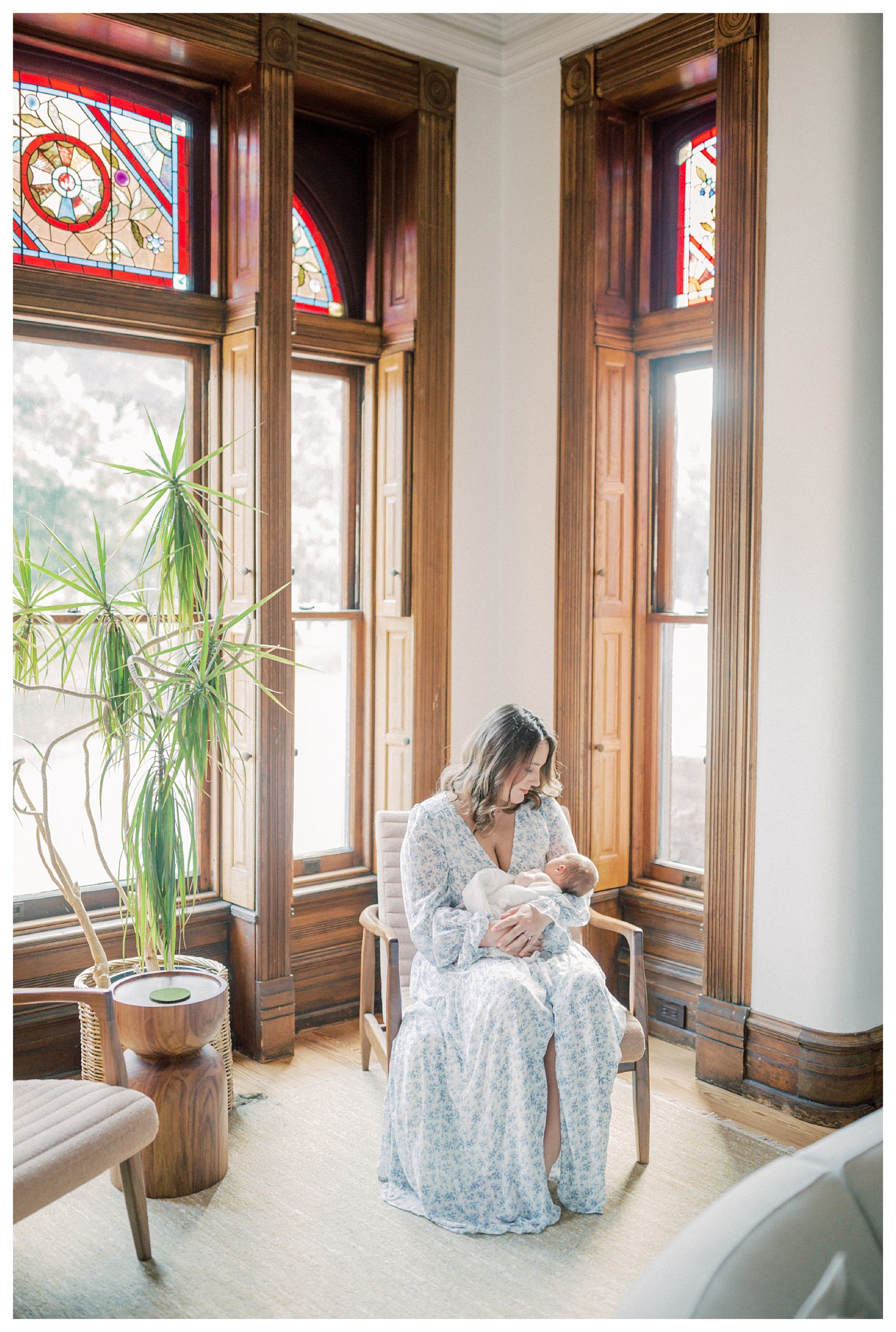 Mother Looks Down At Baby While Sitting In Her Living Room During In-Home Newborn Session