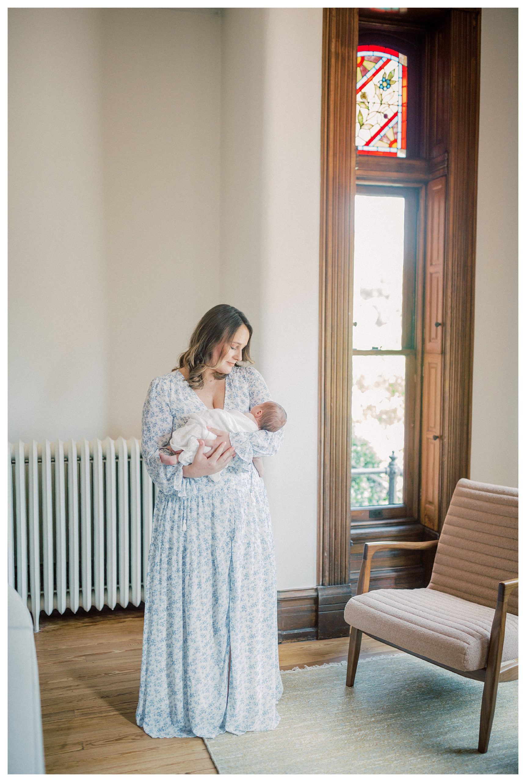 New Mother In Blue Floral Dress Holds Newborn Baby While Standing In Living Room During In-Home Newborn Session