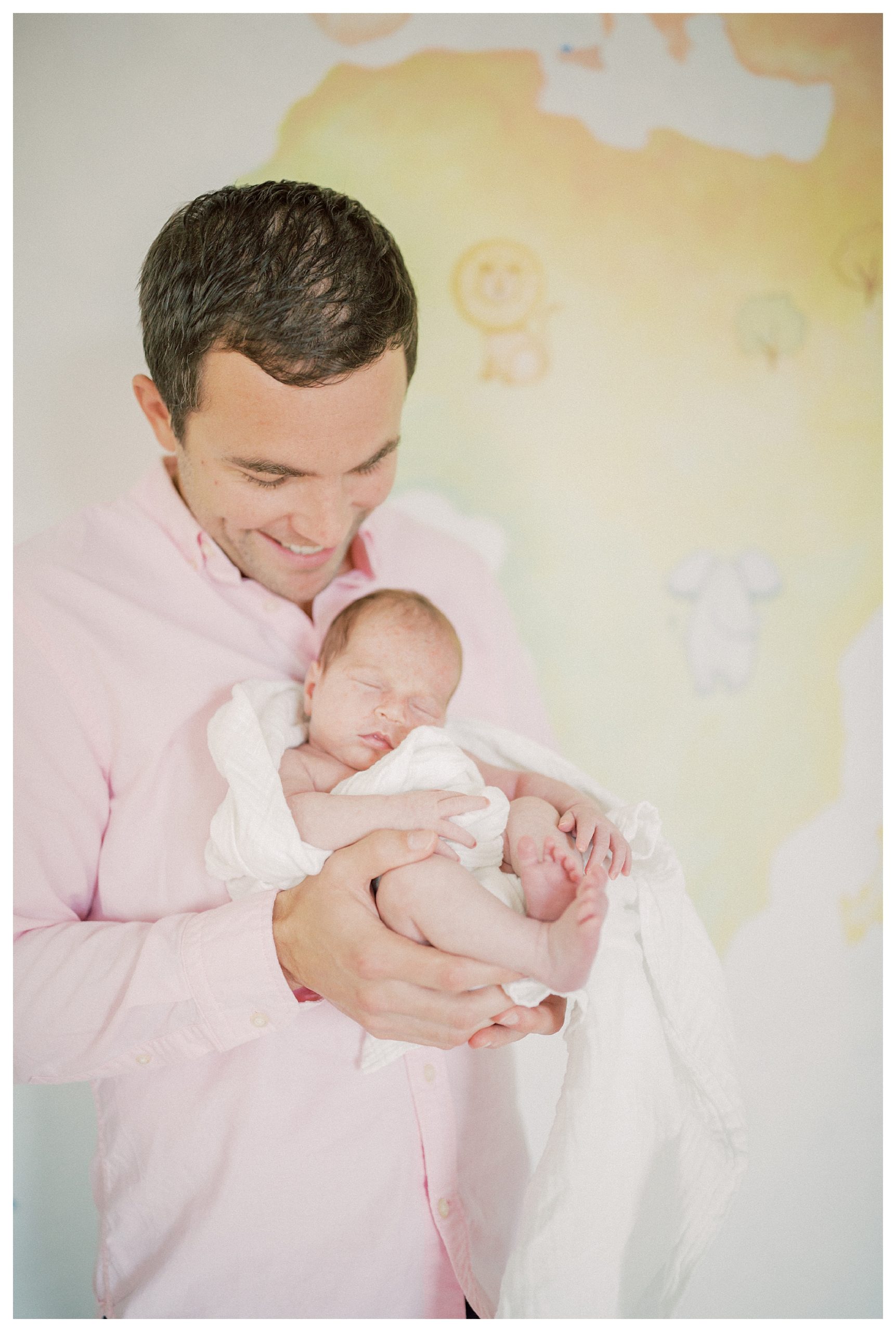 Father Holds Infant Daughter And Smiles At Her While Standing In Front Of Colorful Mural In Nursery.