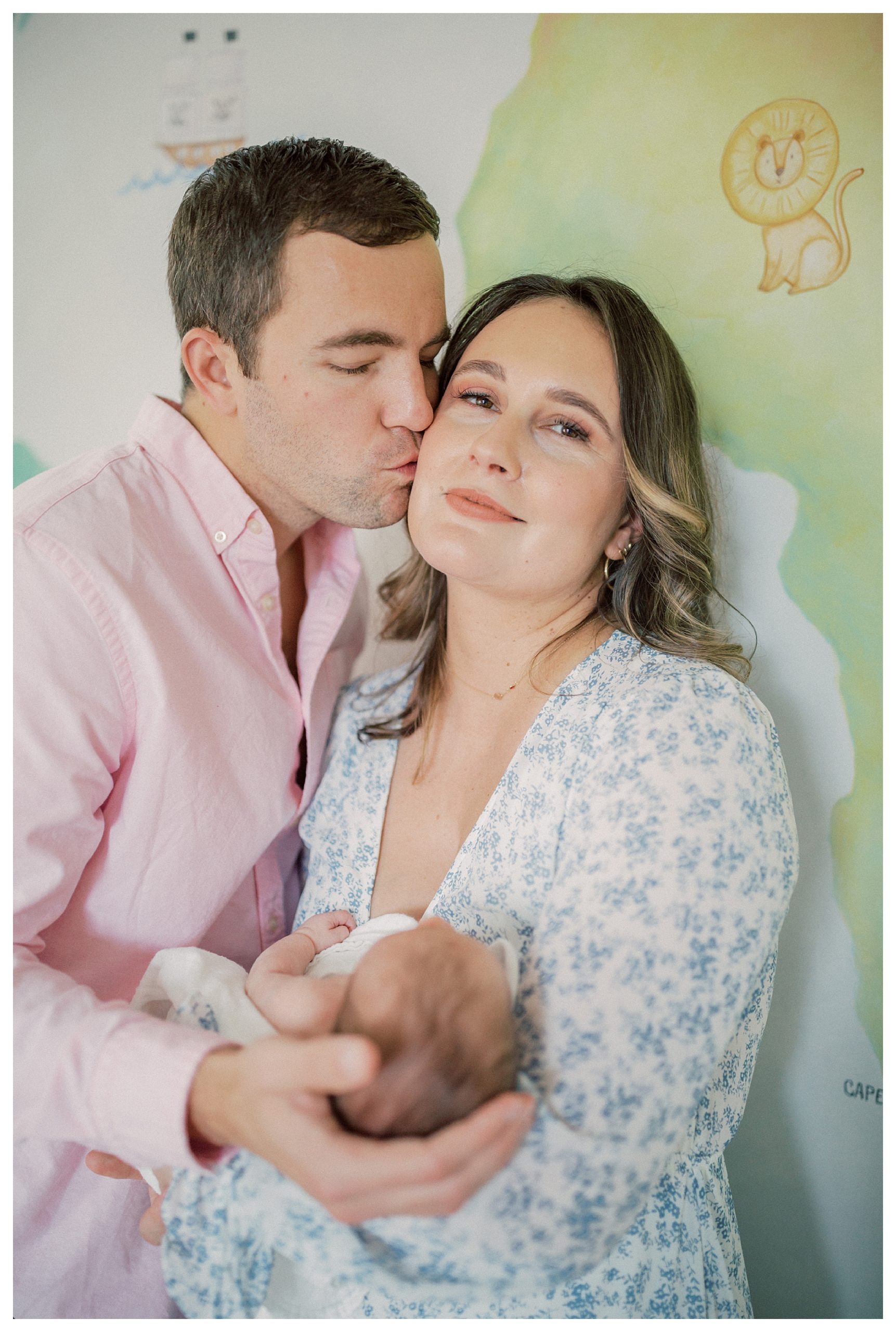 Father Kisses His Wife On The Cheek As She Looks At The Camera Holding Their Newborn Baby.