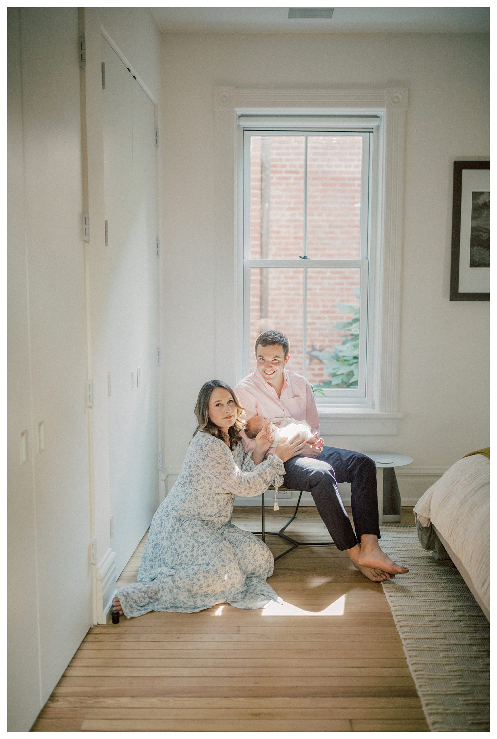 Mother Sits On Floor Touching Her Newborn Baby Held By Her Husband During In-Home Newborn Session.