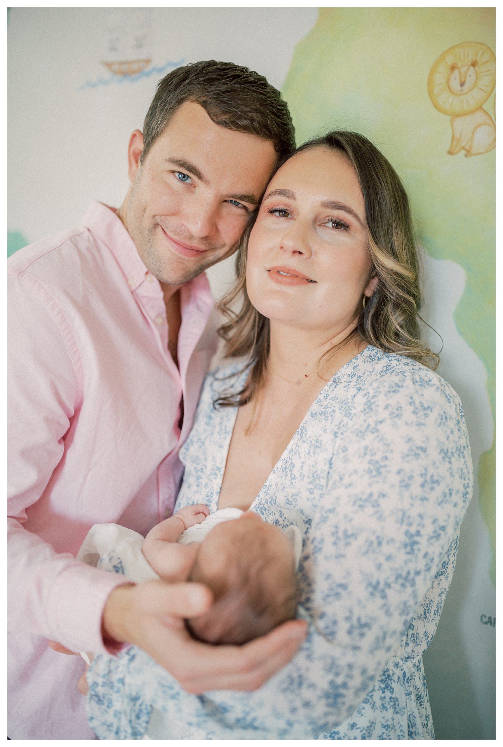 Father And Mother Stand Together Looking At The Camera While Holding Their Newborn Baby During In-Home Newborn Session.