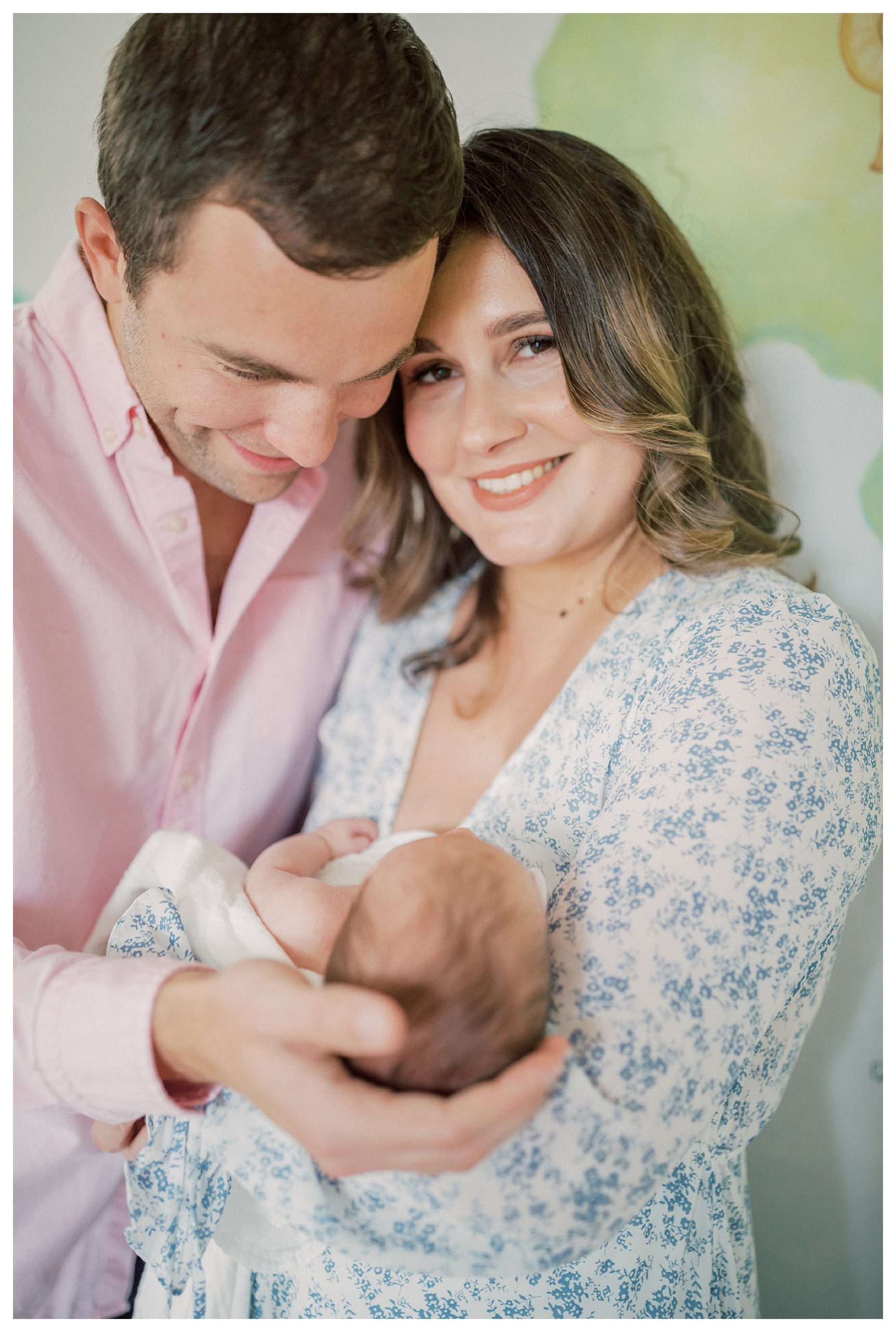 Mother Looks At The Camera And Smiles As Her Husband Leans Over Looking And Smiling At Newborn Baby.