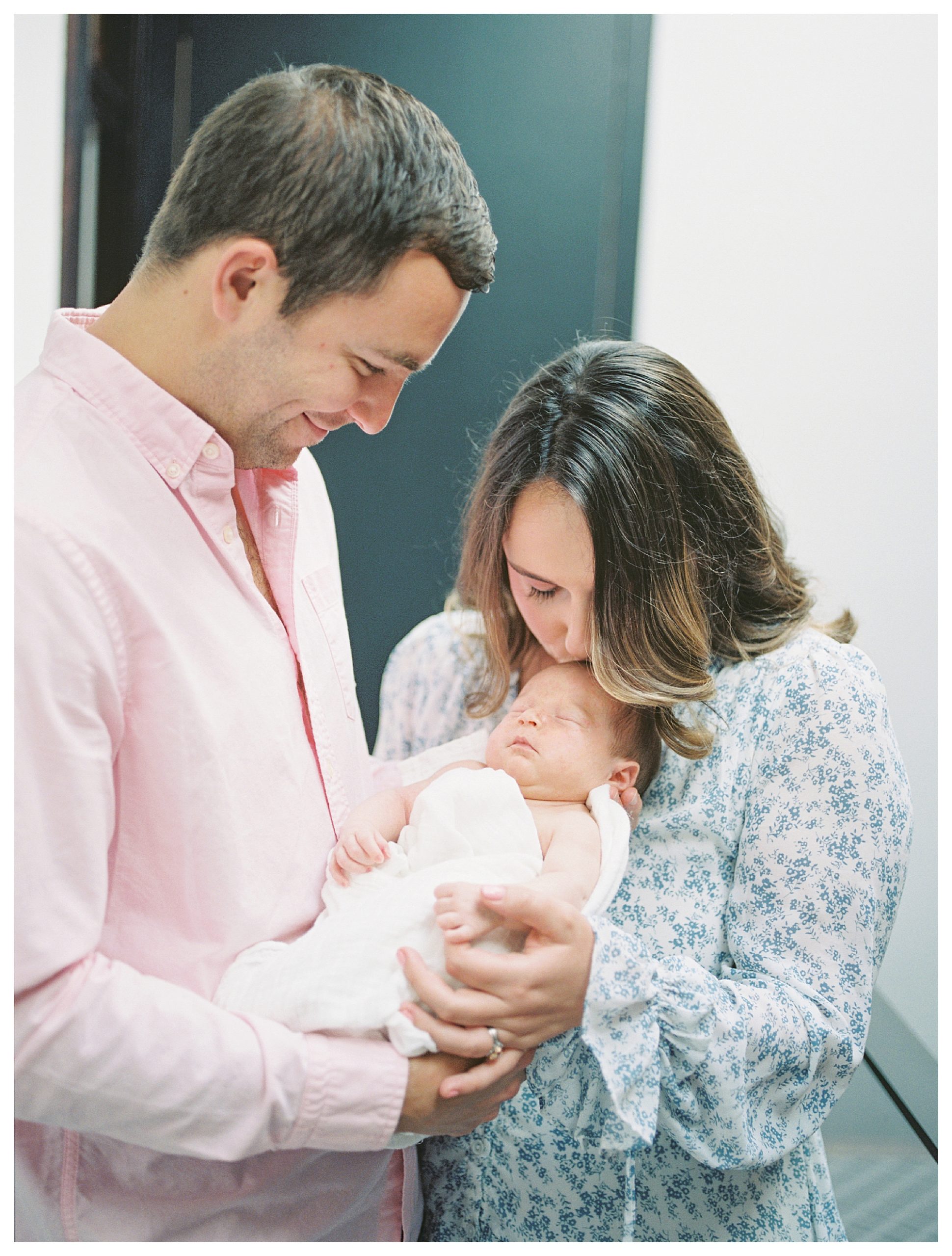 Mother In Blue Floral Dress Leans Over To Kiss Baby's Head While Baby Is Held By Father During In-Home Newborn Session.