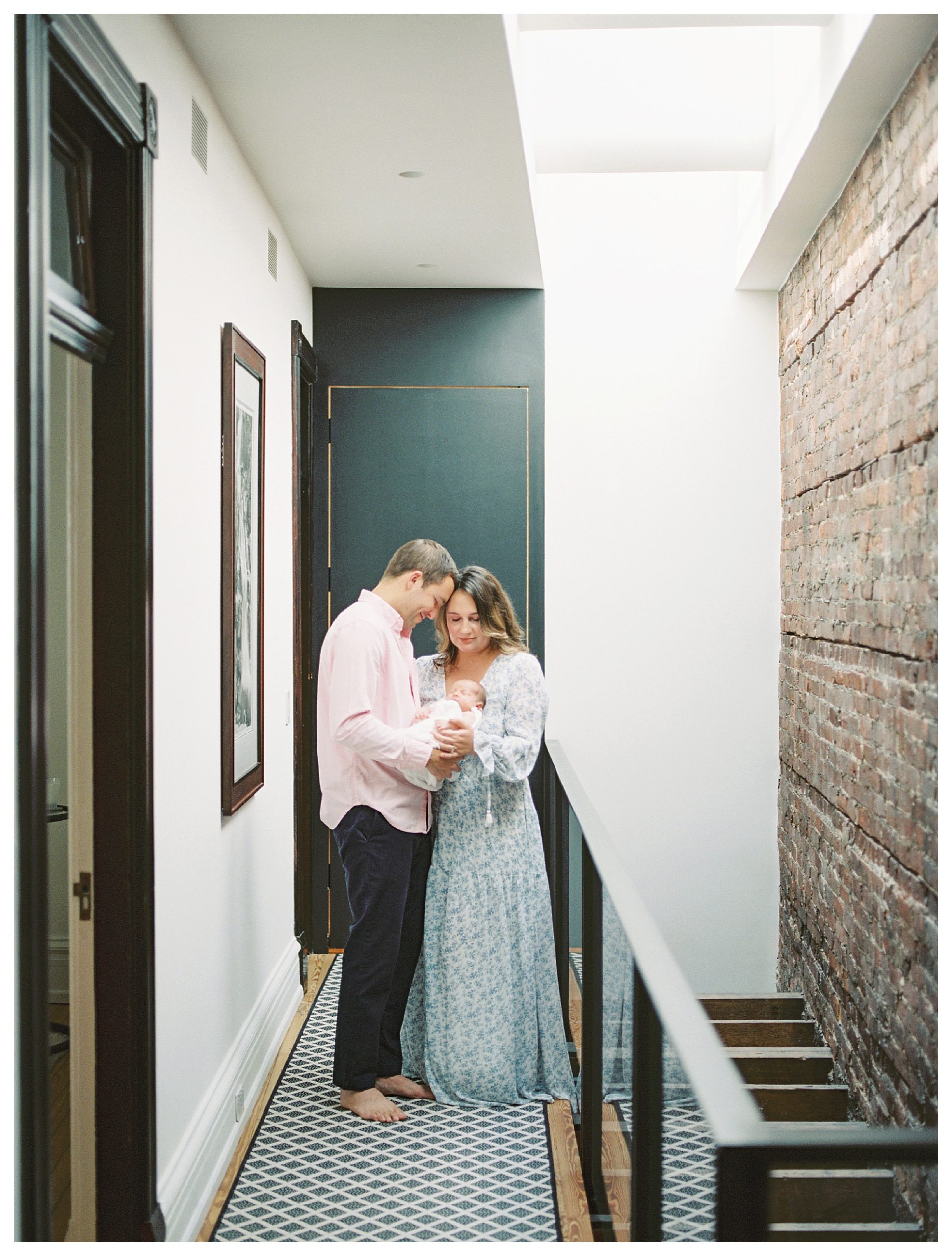 New Parents Stand In Their Hallway, Leaning Into One Another Holding Their Newborn Baby During Their In-Home Newborn Session.