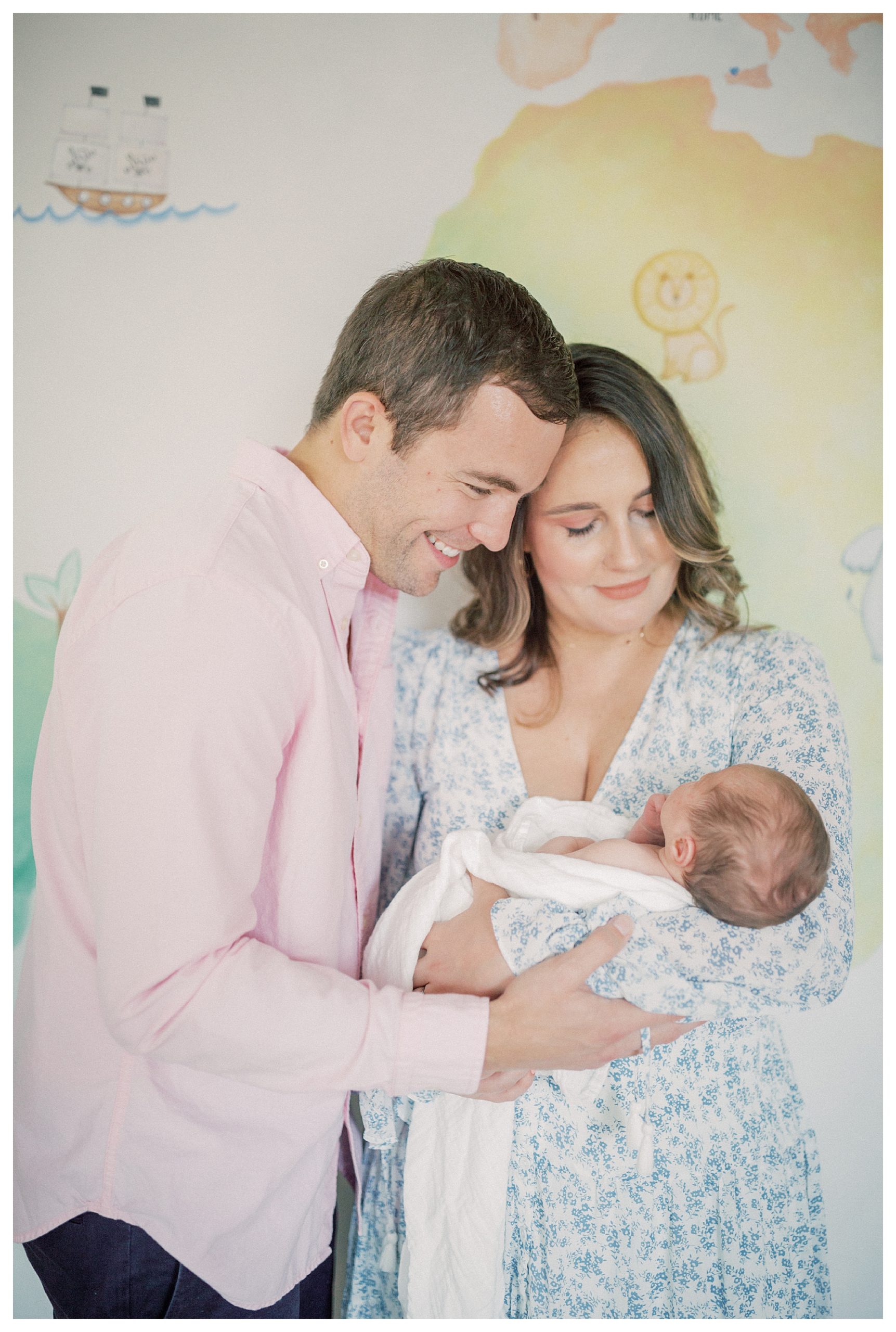 Father And Mother Stand In Front Of Colorful Mural In Nursery, Holding Newborn Baby