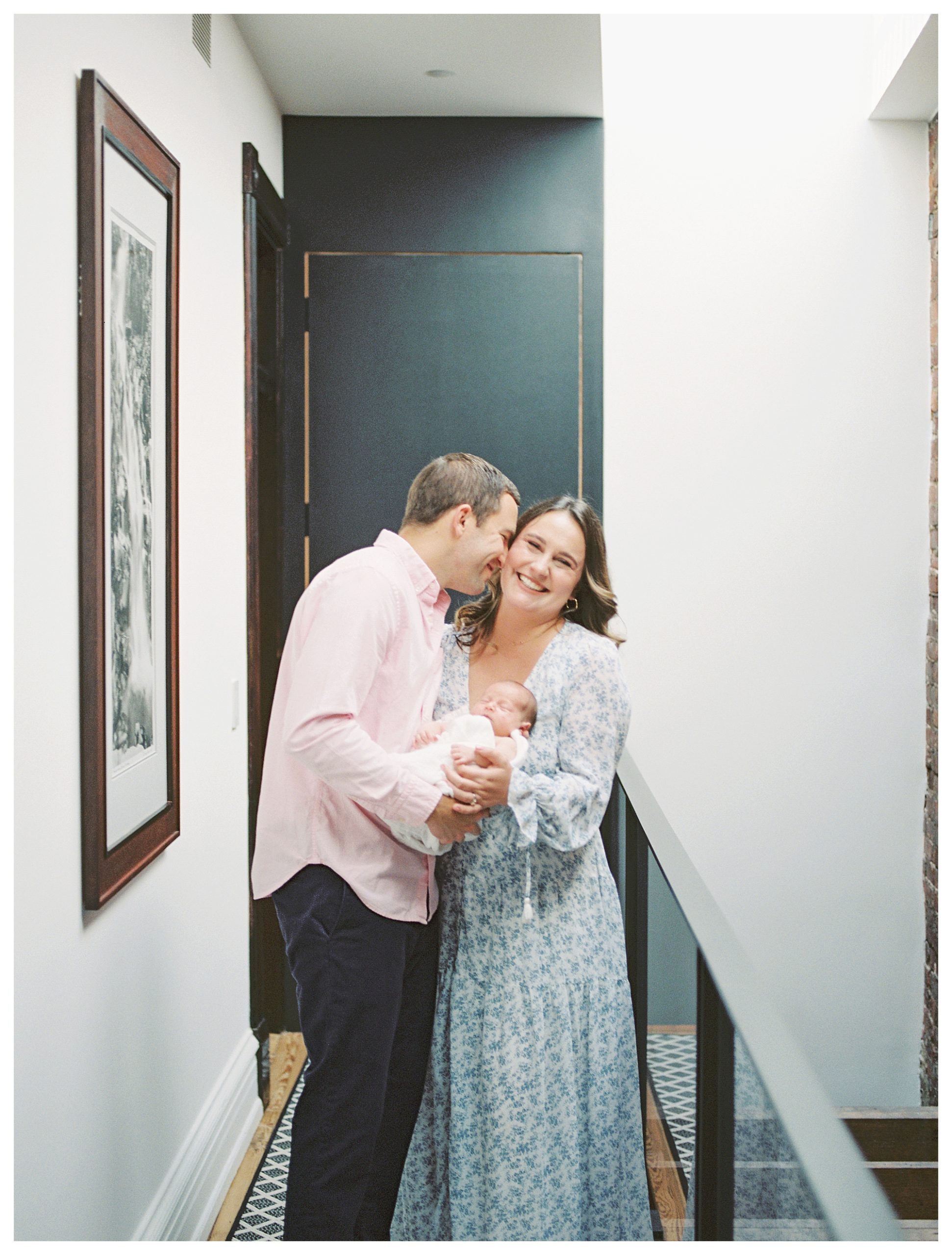 Father Leans Into His Wife And Nuzzles Her As They Hold Their Newborn Baby During Their In-Home Newborn Session.