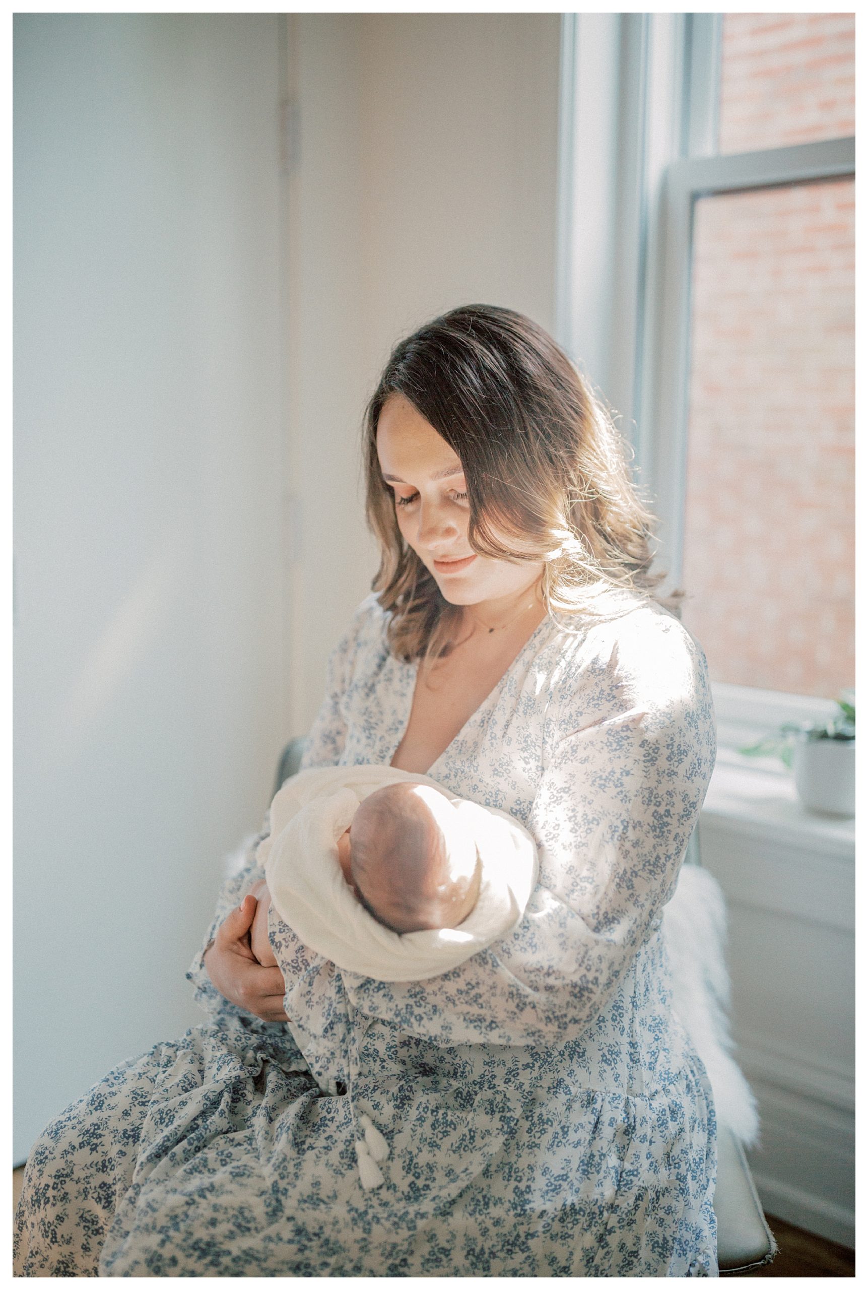 Mother Looks Down At Her Newborn And Smiles As She Sits In A Chair In The Light During Their In-Home Newborn Session.