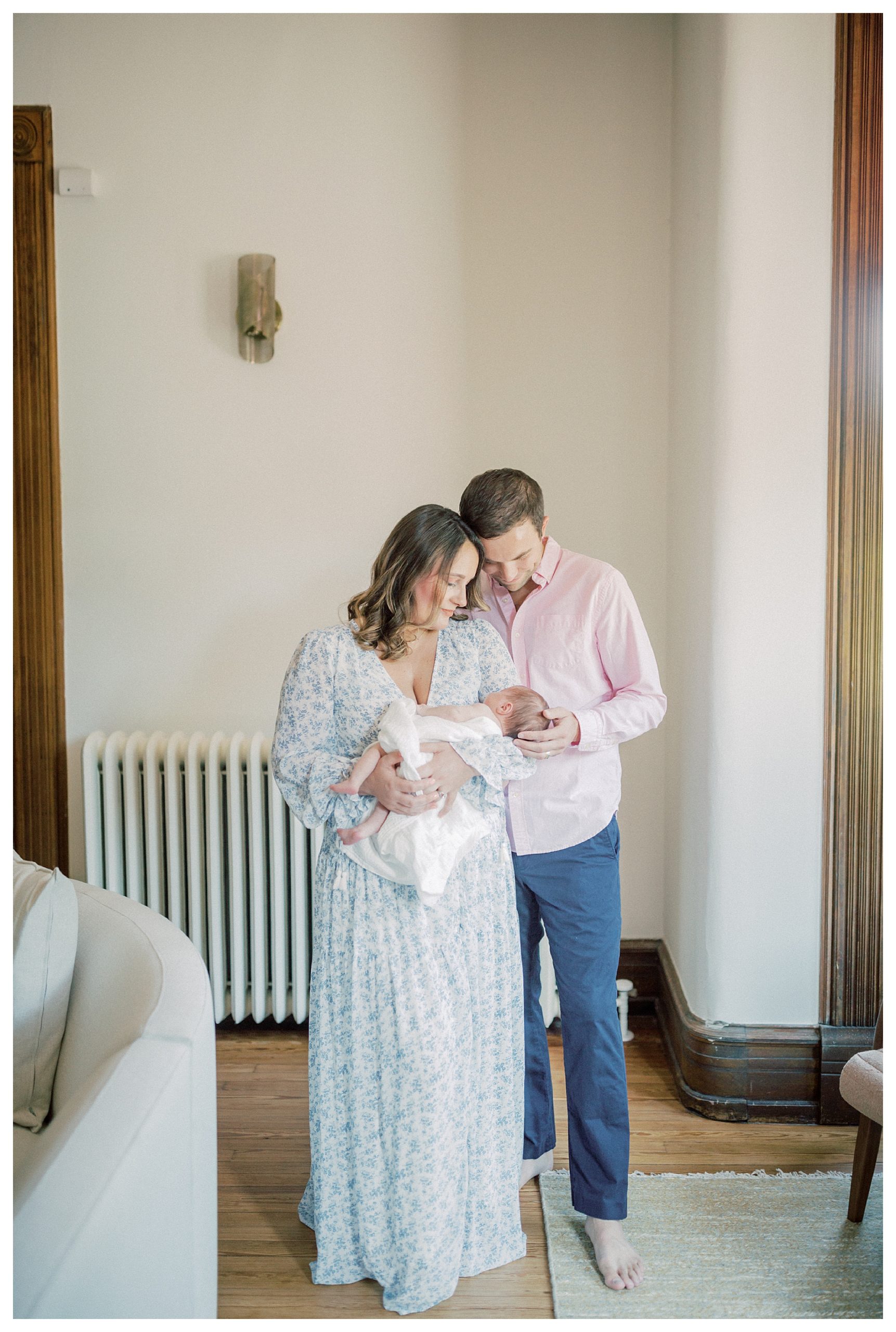 Mother And Father Hold Newborn Baby And Stand Close To One Another During Their In-Home Newborn Session.