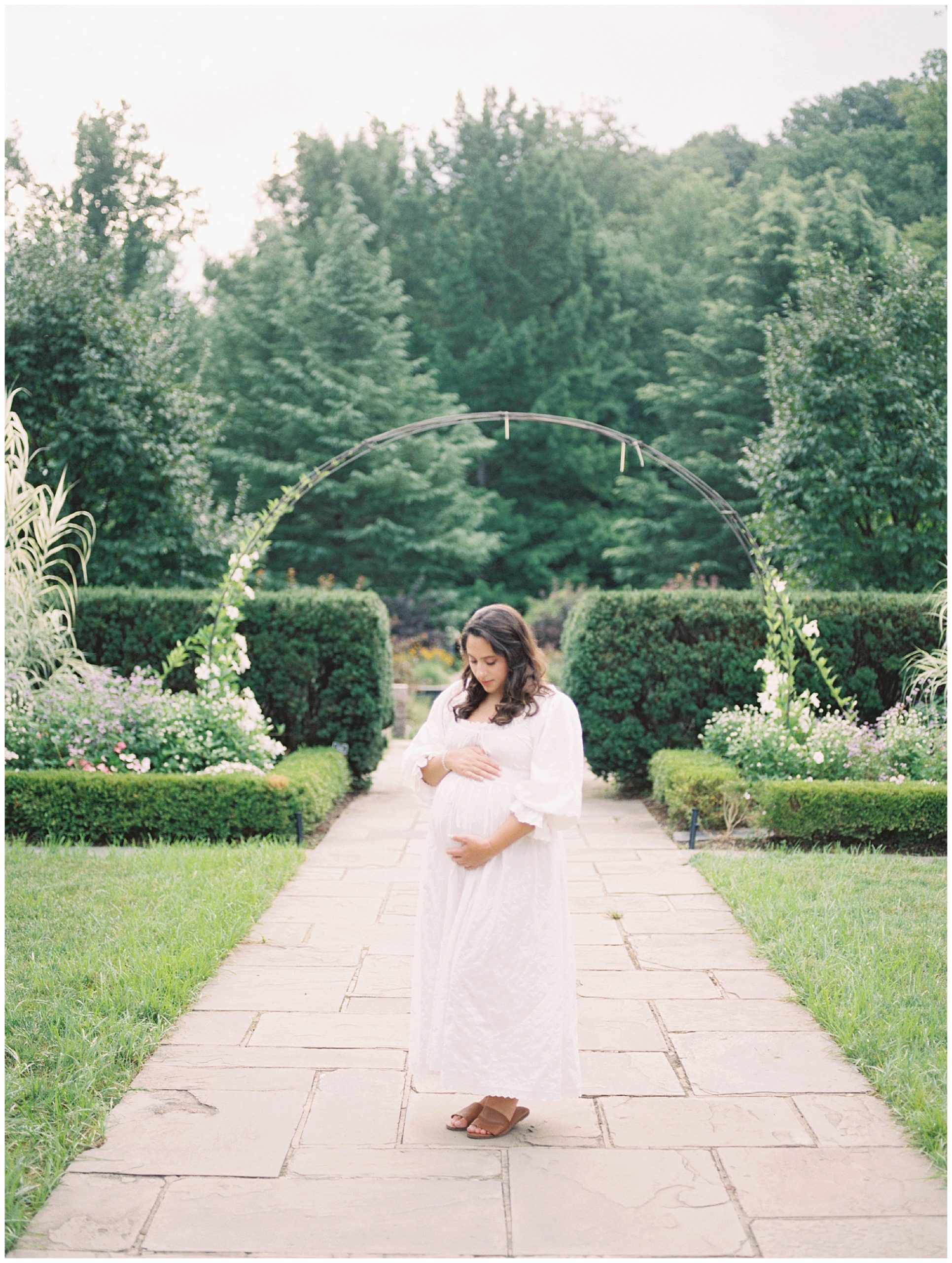 Pregnant Mother In White Doen Dress Stands With One Hand On Top And One Hand Below Her Belly By A Floral Arch In Brookside Gardens.