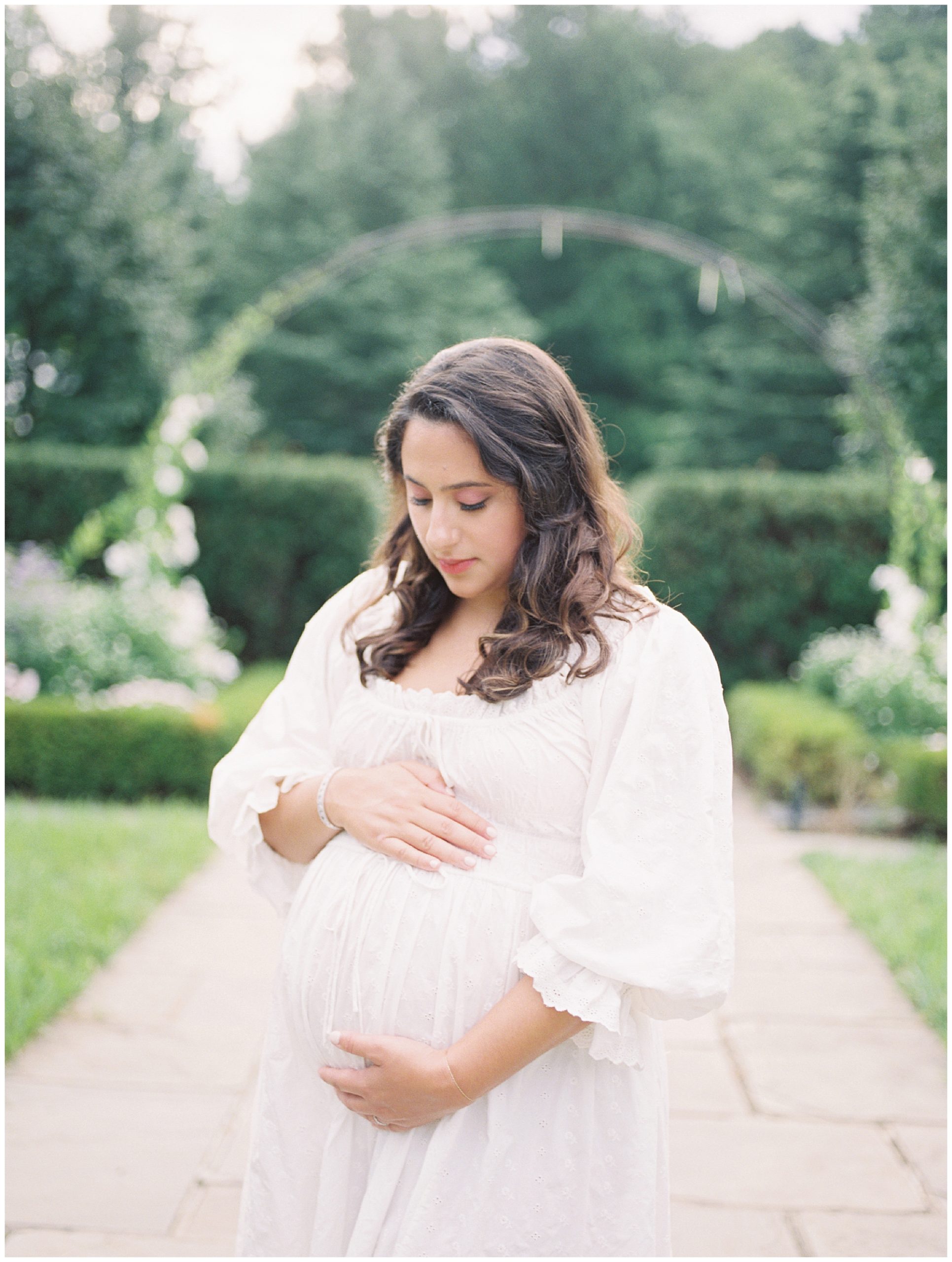 Expecting Mother In White Doen Ischia Dress With One Hand On Top And One Hand Below Her Belly, Looking Down.