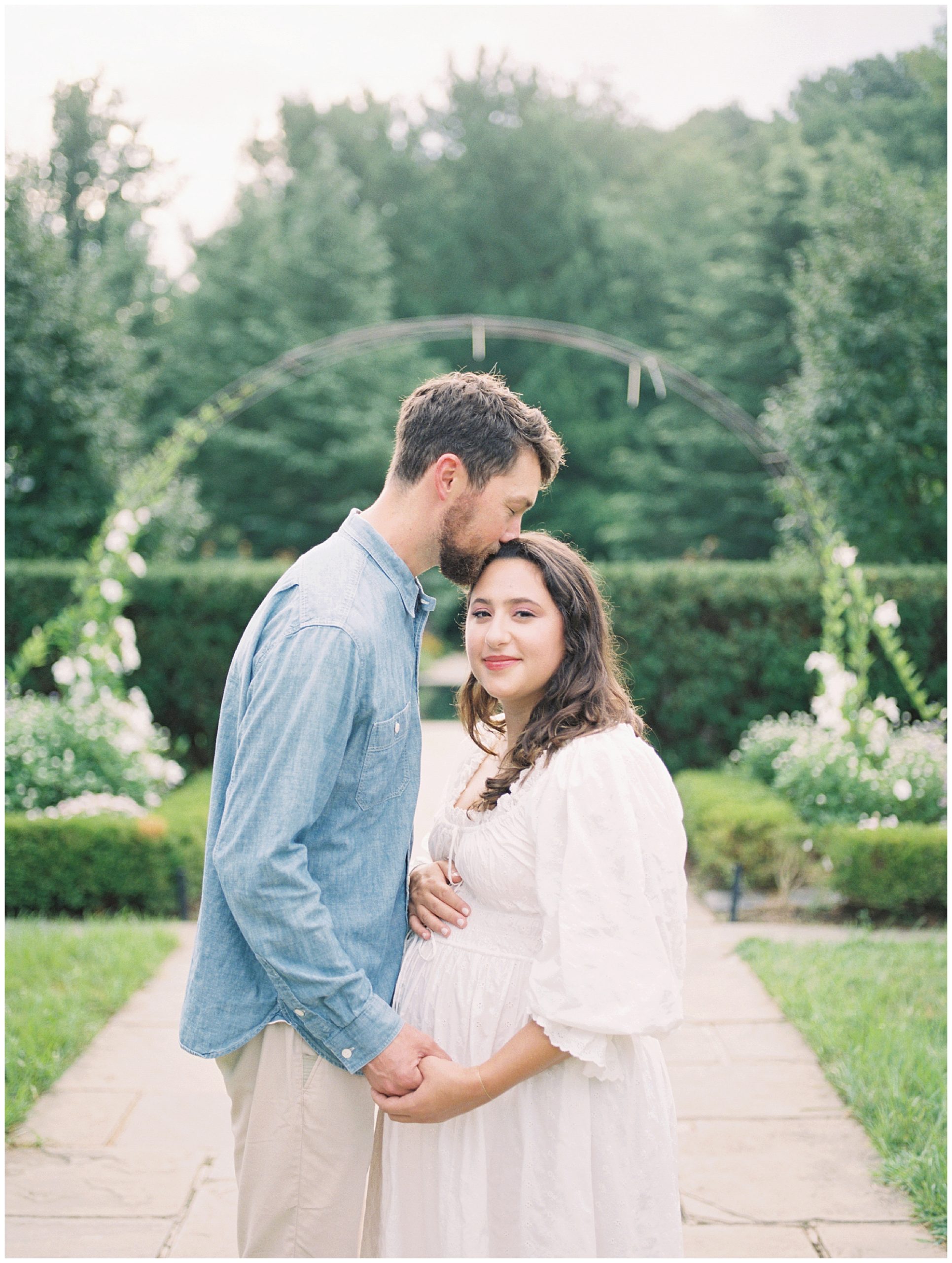 Father Kisses His Wife's Forehead As She Looks At The Camera During Their Maternity Session At Brookside Gardens.